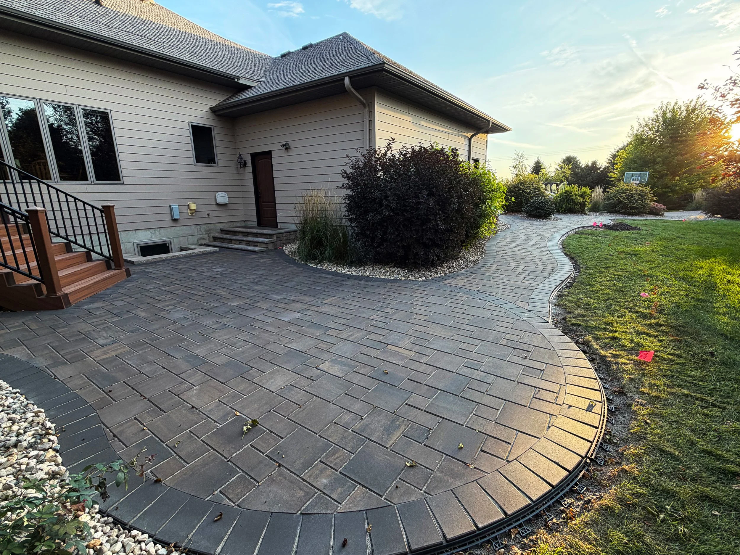 A freshly paved, curved stone patio in the backyard of a house, with steps leading to a door and a landscaped garden area with bushes and trees, and a sunset in the background.