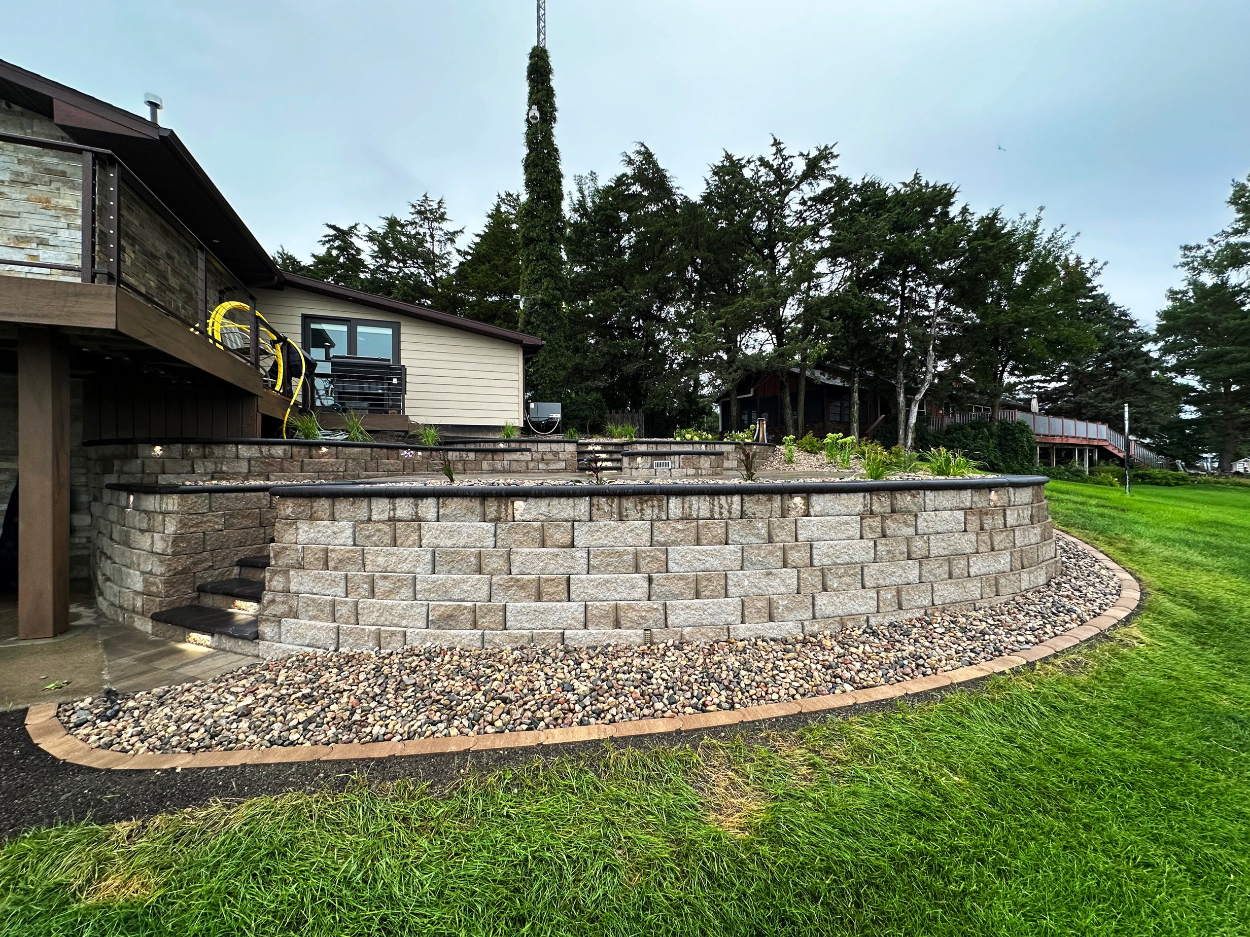 A multi-level backyard with stone retaining walls, a green lawn, trees, and outdoor structures.