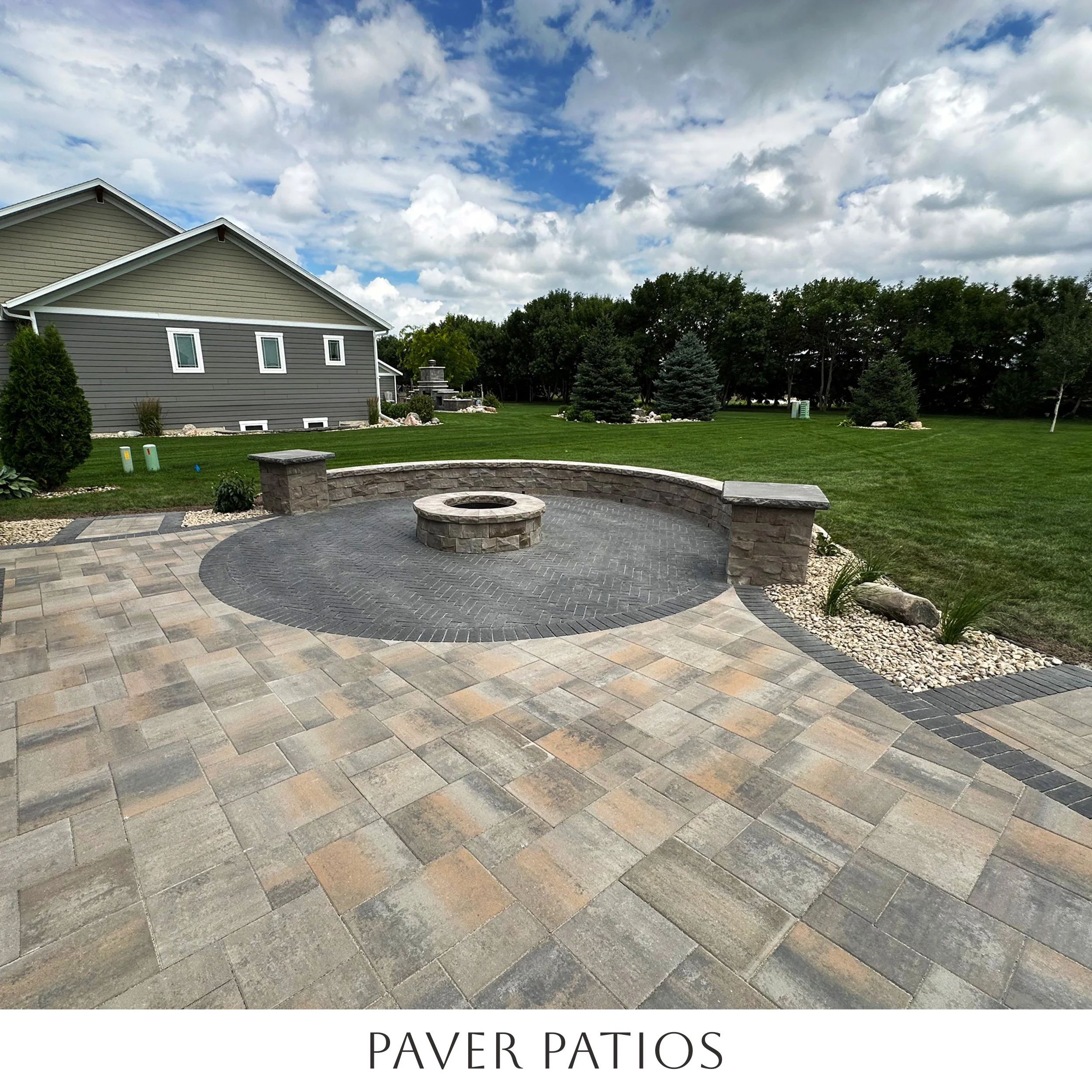 View of a backyard paver patio with a fire pit, surrounded by a curved stone wall. The patio has a varied stone pattern, with a grassy lawn and trees in the background, and a partly cloudy sky overhead.