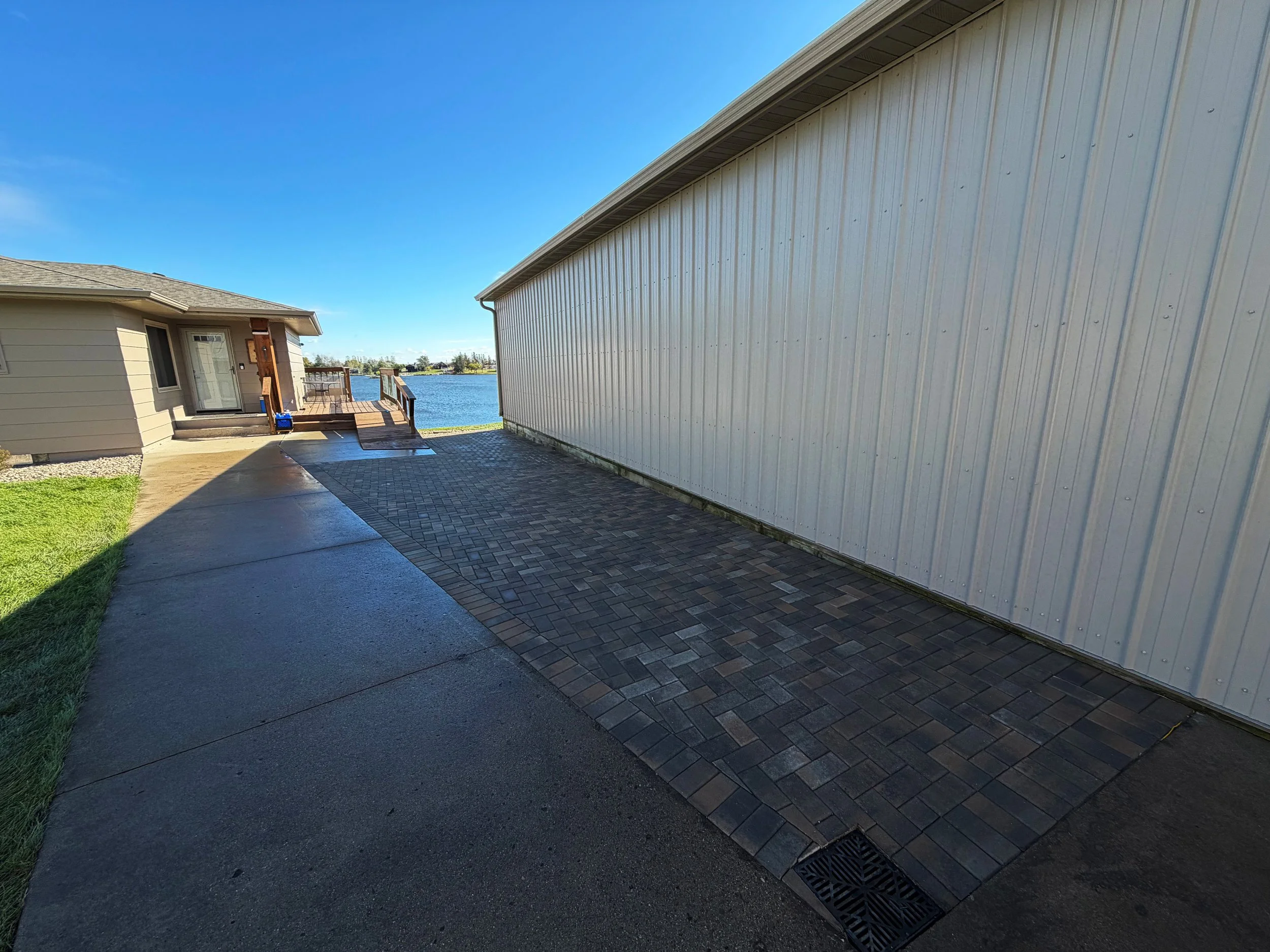 Backyard with a sliding glass door leading to a small wooden deck overlooking a body of water, with a paved area with different paving materials adjacent to the house and a grassy area on the left. The house front features horizontal siding, and the 