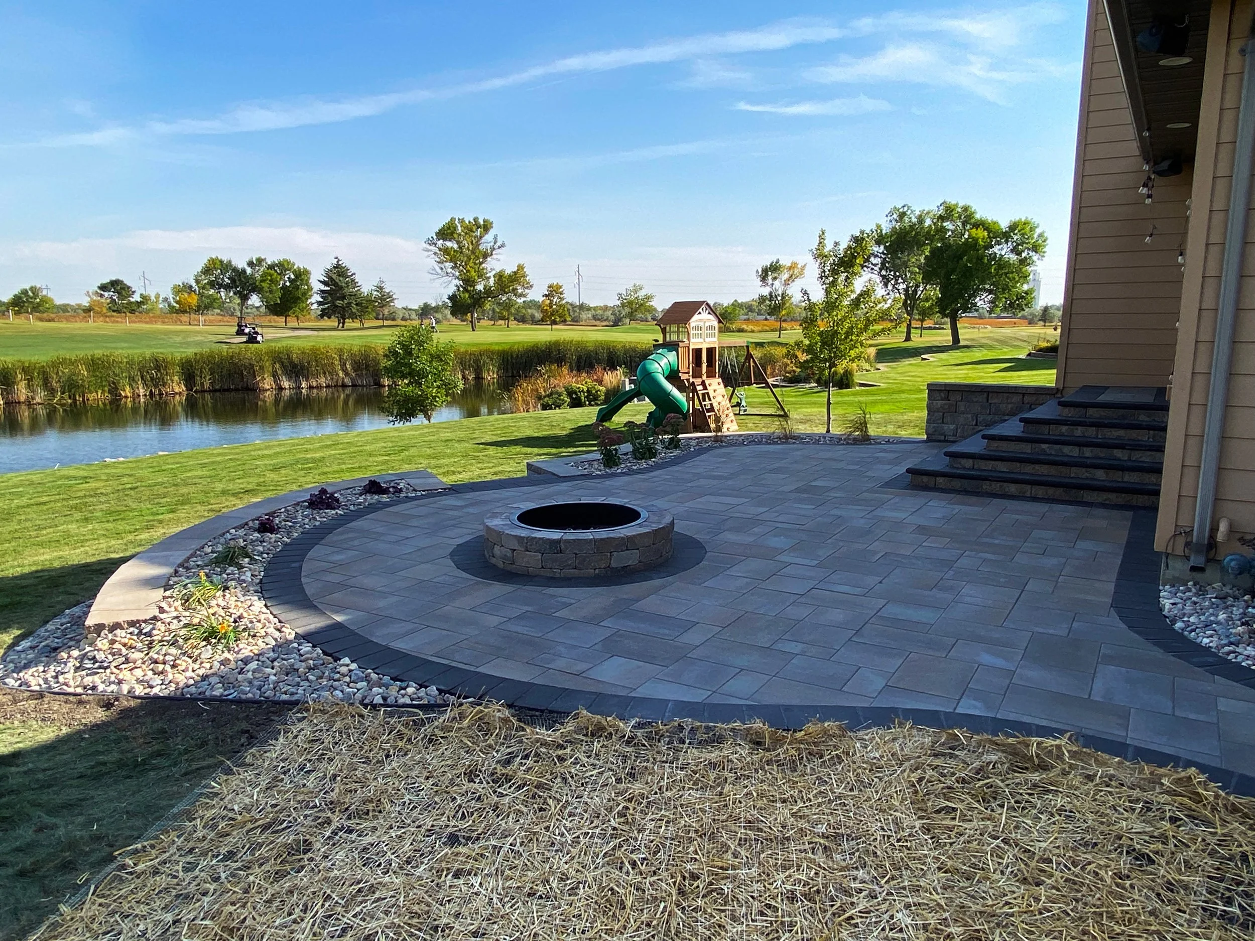 Backyard patio with stone pavers, a fire pit, and a children's playset with a slide near a pond and open grassy area.
