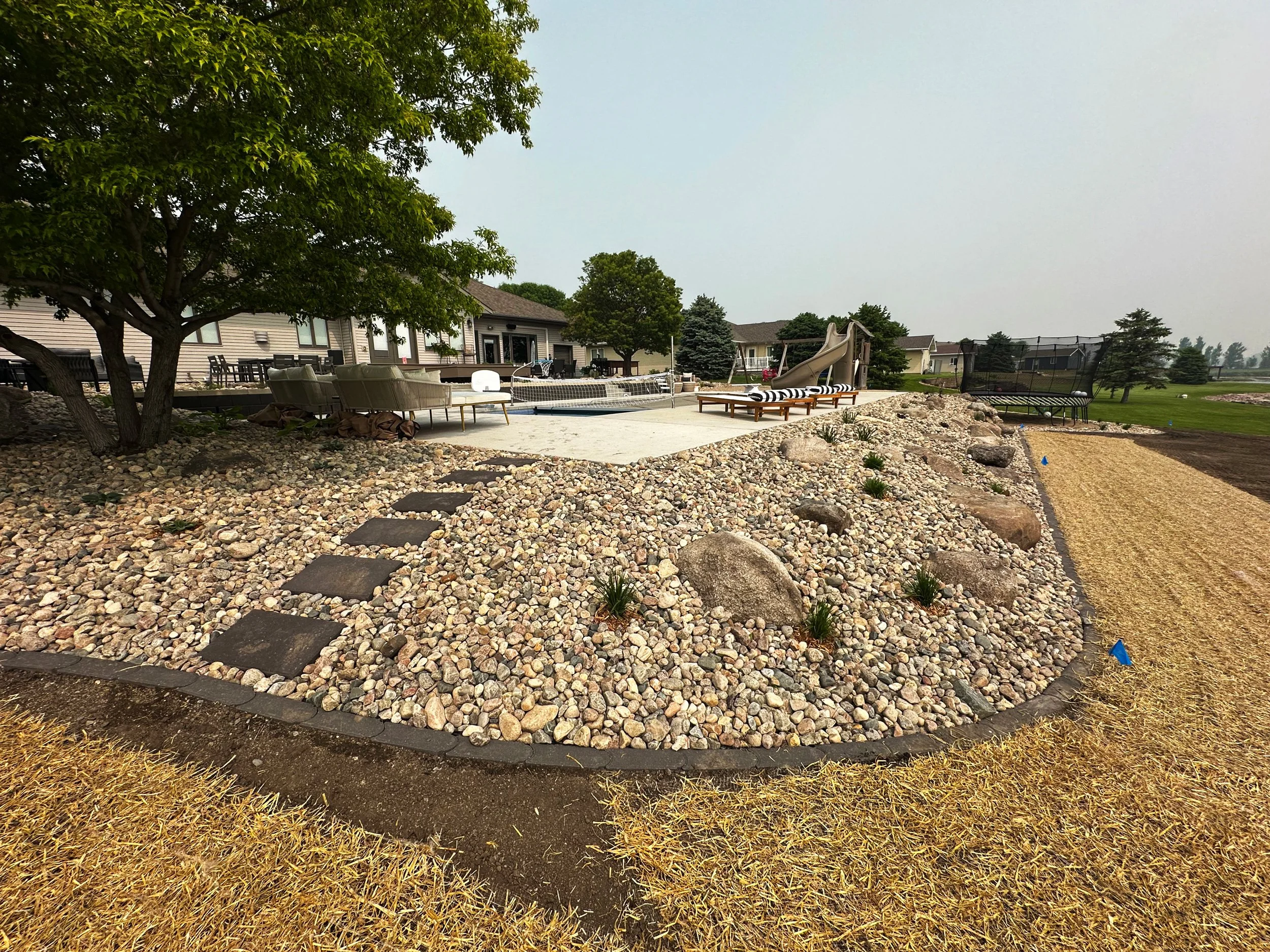 Backyard with a stone pathway, a tree on the left, a patio with furniture and a slide, a trampoline, and open grassy area in the background.