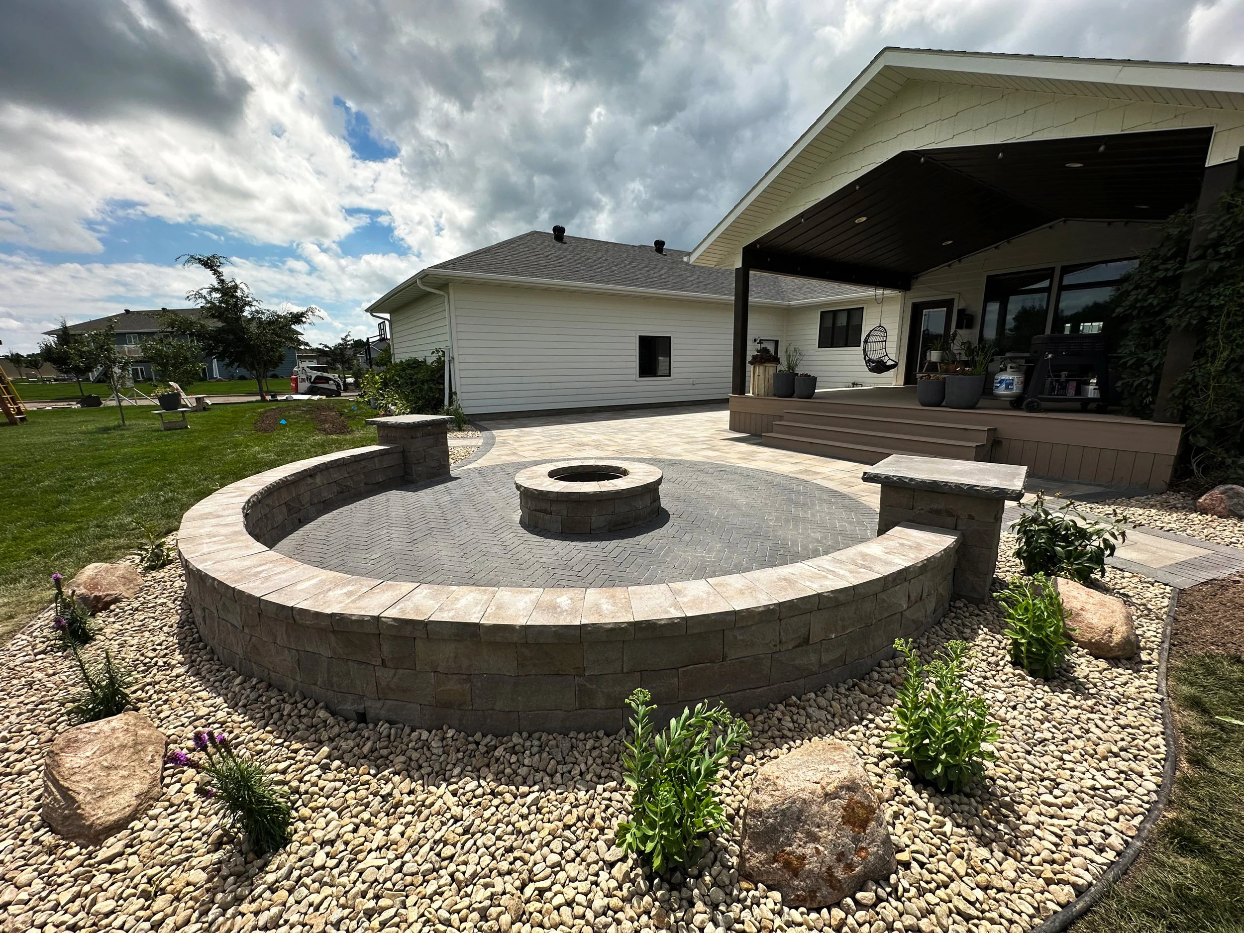 Backyard patio with circular stone fire pit and retaining wall, surrounded by small plants and rocks. There is a house with a porch and stairs leading down to the patio, with outdoor furniture and equipment visible on the porch. The area is landscape