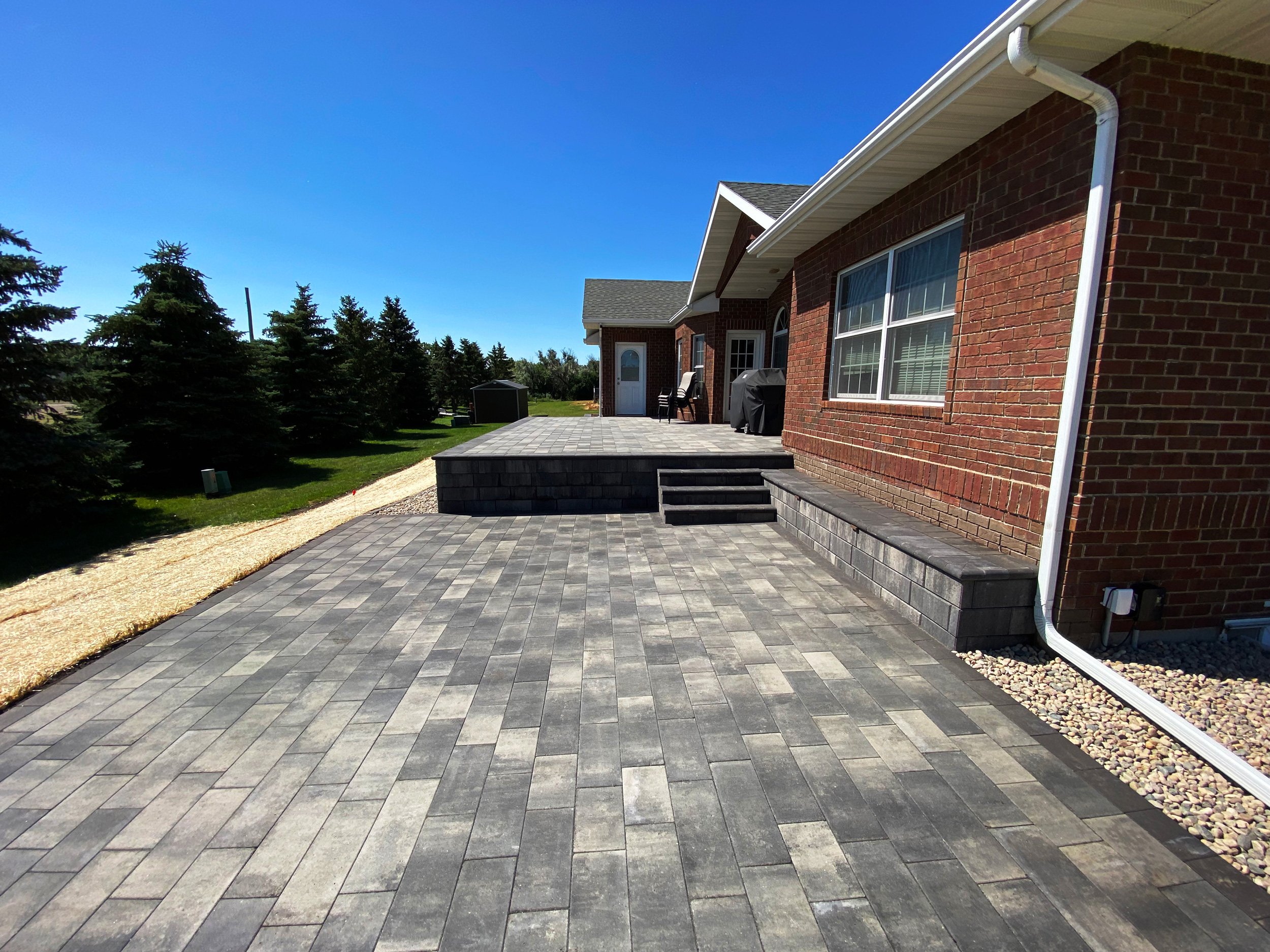 Backyard patio area with gray brick pavers and a raised section with steps, attached to a red brick house with a large window and white trim, on a sunny day with a clear blue sky.