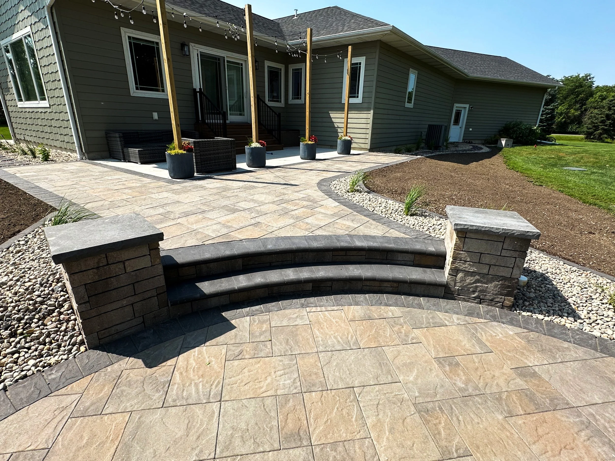 Backyard patio with a small set of three black curved steps leading to a stone-paved patio area. The patio features a grey house with a small deck, sliding glass door, and outdoor seating. There are potted plants with flowers along the deck, string l