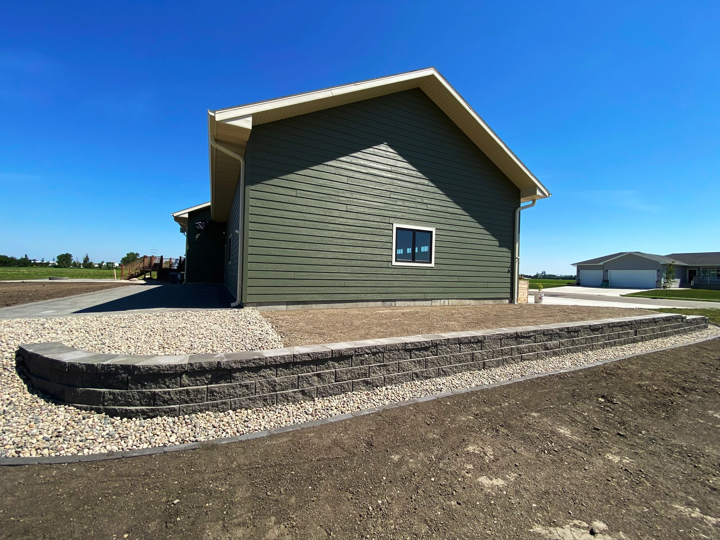 Side view of a green house with a sloped roof, new landscaping with a curved stone retaining wall, gravel, and freshly tilled soil, under a clear blue sky.