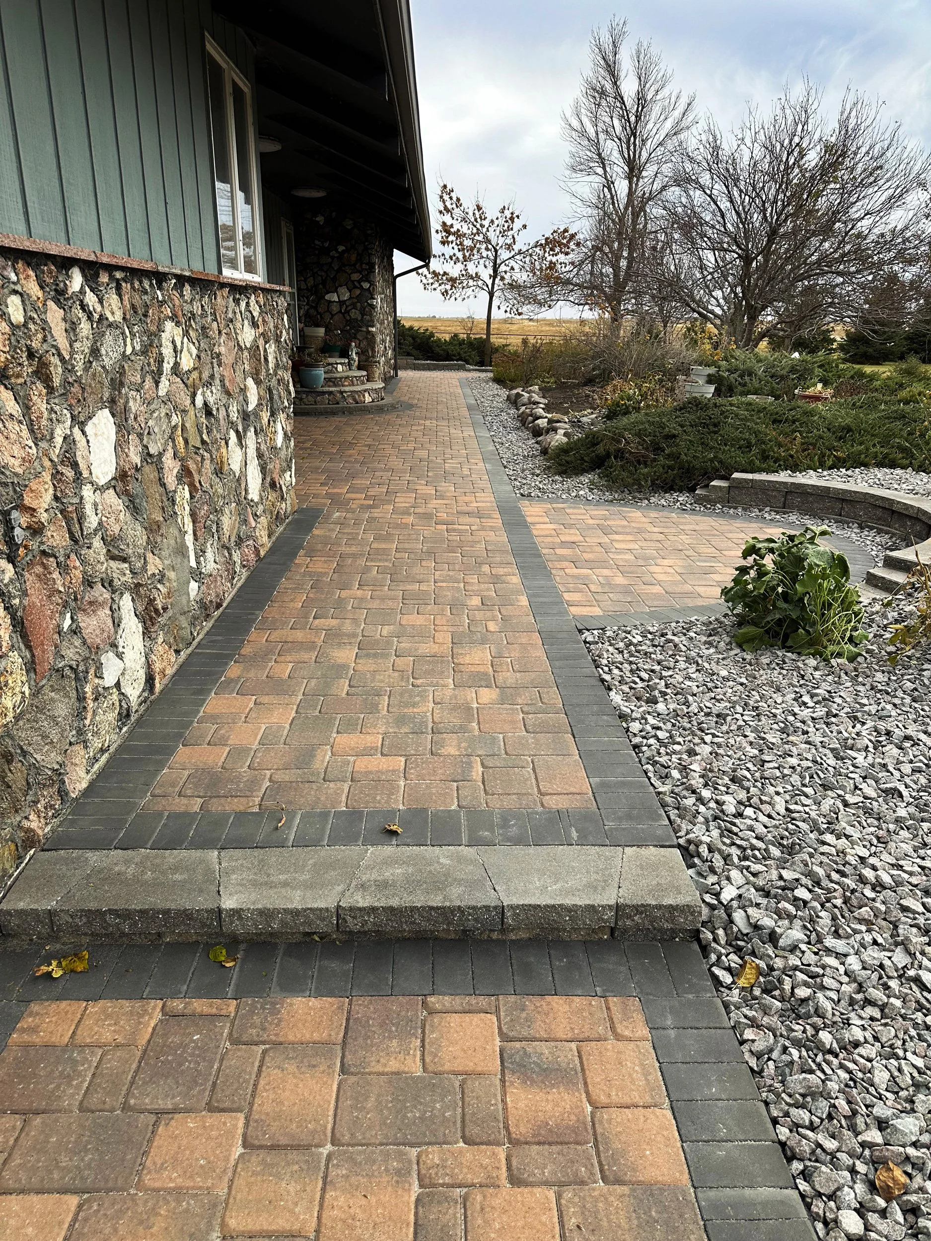 Paved walkway with bricks and stones along a house with a stone and green siding exterior, surrounded by a landscaped yard with trees and plants.