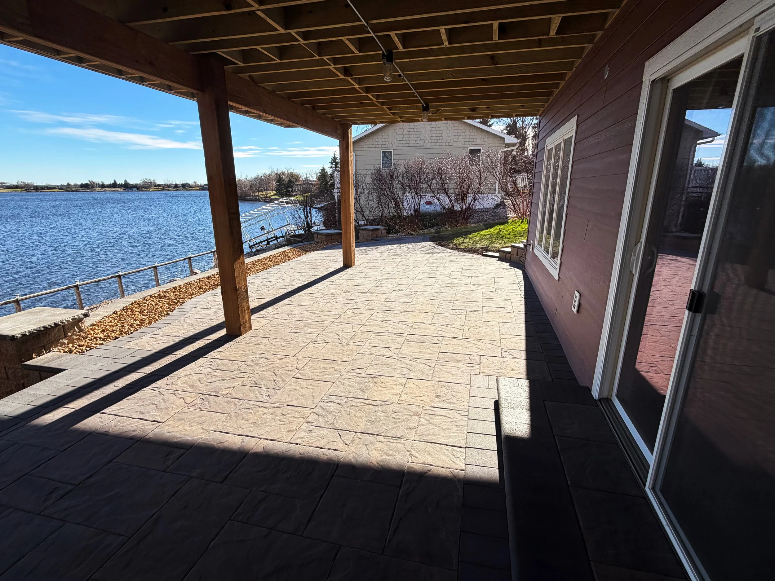 Covered patio with stone flooring overlooking a body of water, with a house and trees in the background.