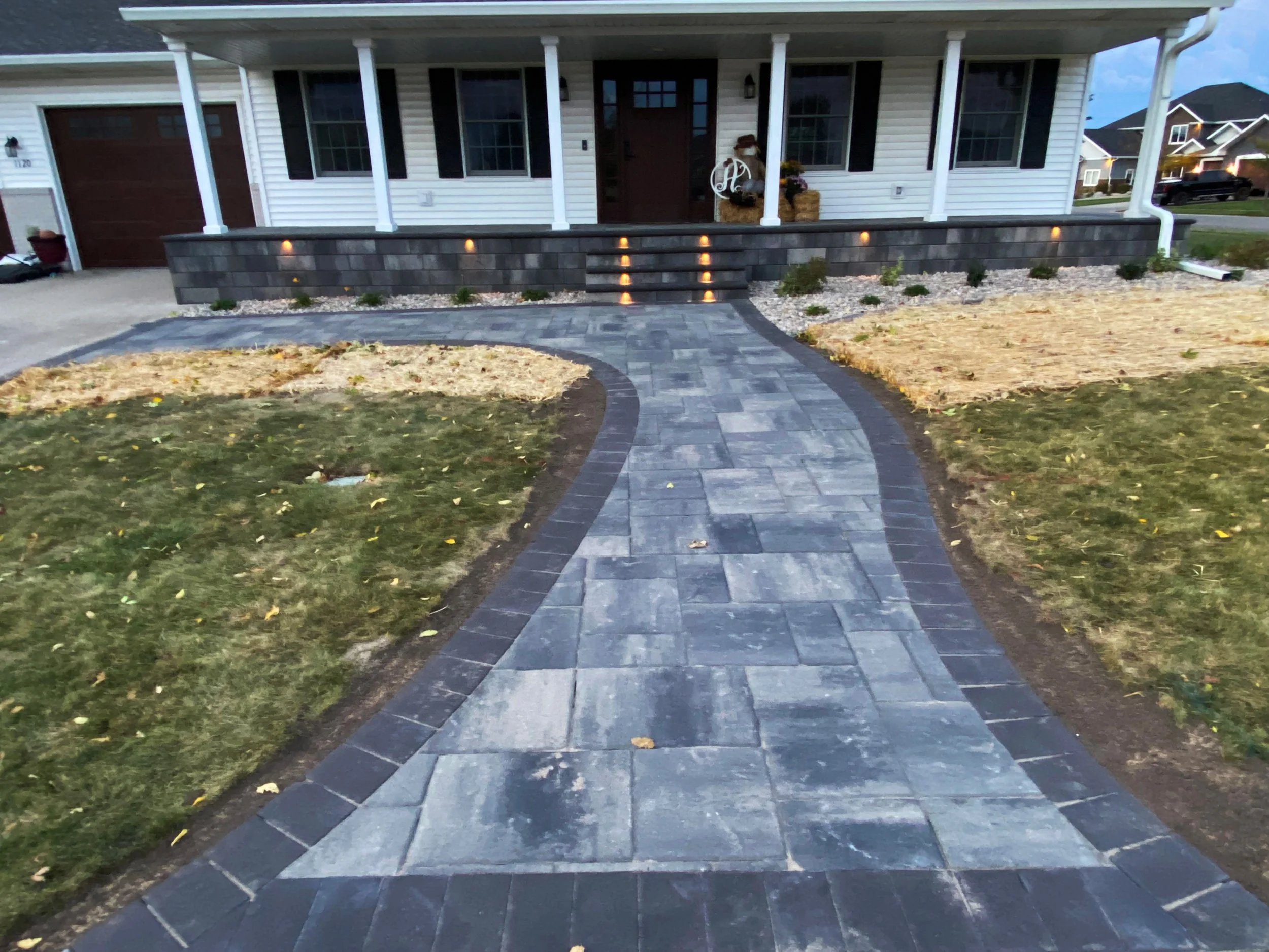 Curved stone pathway leading to a front porch with steps, landscaping, and outdoor lights, in a suburban neighborhood.