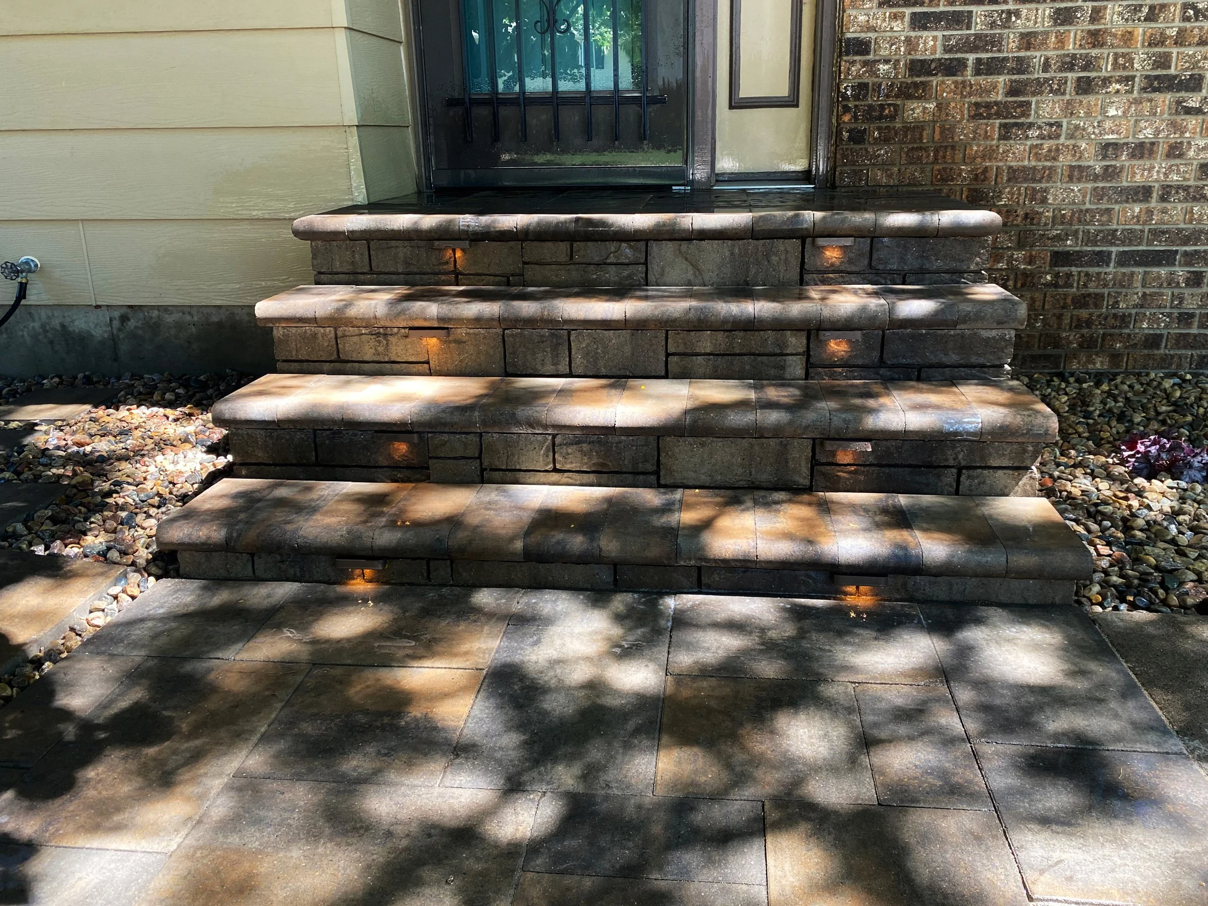 Stone steps leading up to a front door with a screen door, surrounded by a brick and siding exterior, with shadows from trees cast on the steps and sidewalk.