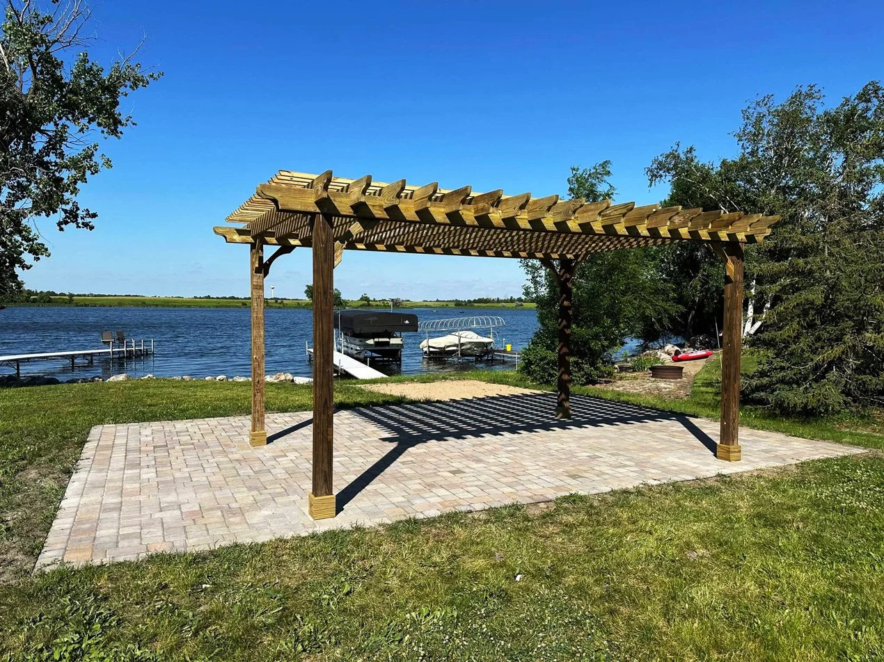 A wooden pergola over a paved patio area near a lake, with a dock, boats, and trees in the background under a clear blue sky.