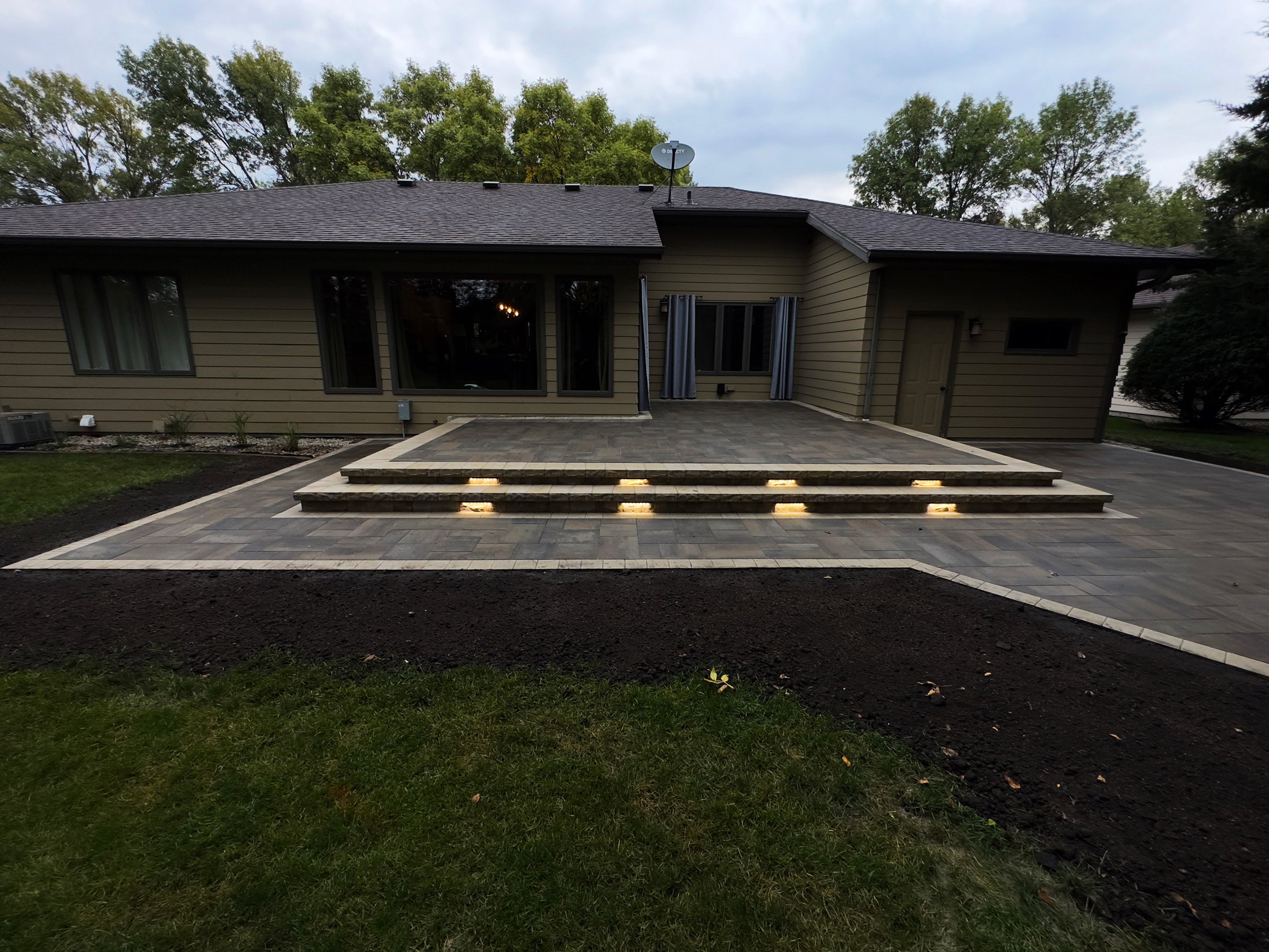 A newly paved backyard patio with three steps leading up to a house with large windows and a sliding door, surrounded by a landscaped yard with green grass and trees in the background.