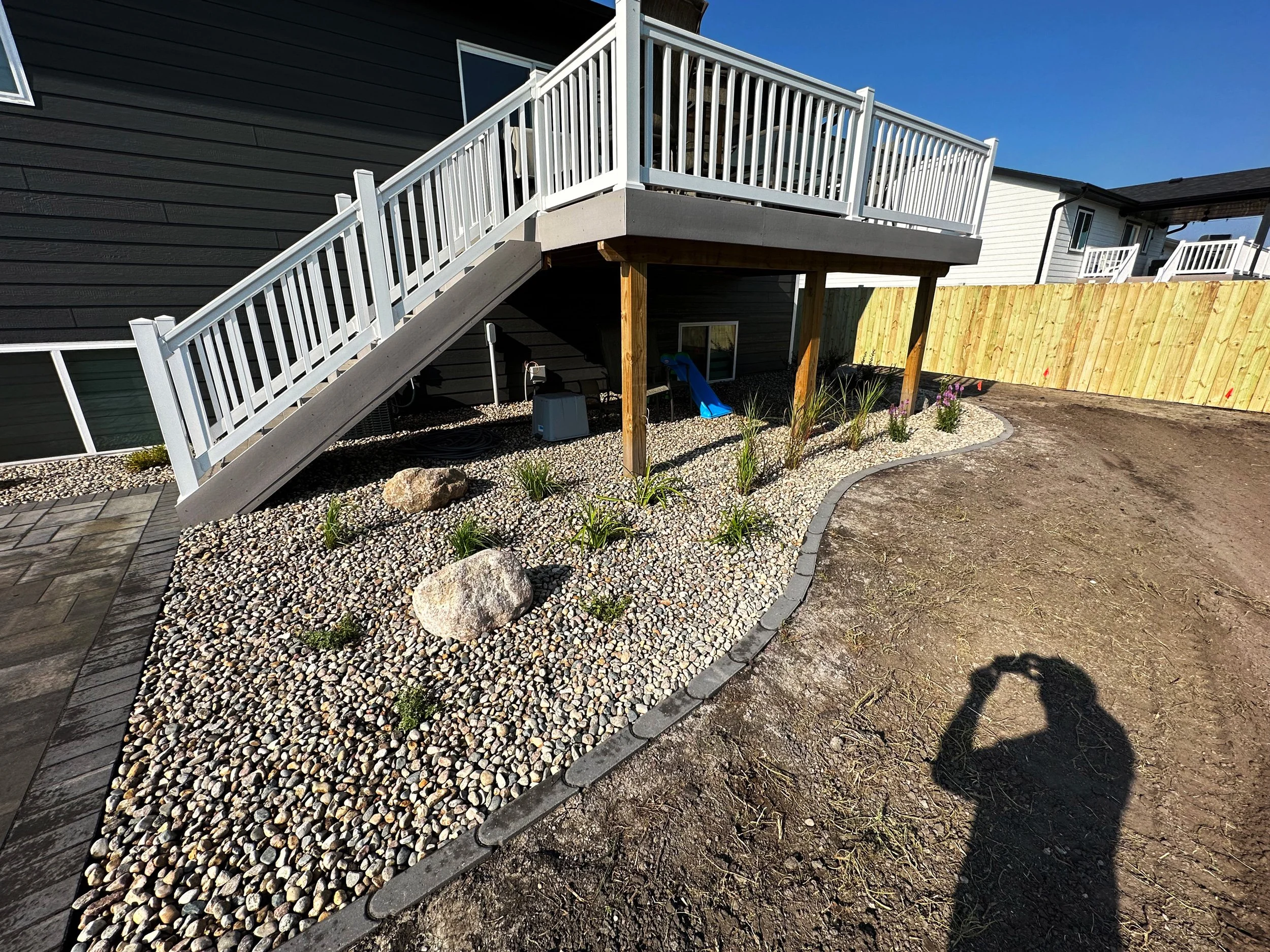 Backyard with a white-railed raised deck, gravel landscaped area with large rocks and plants, and a dirt yard, with neighboring houses visible in the background.