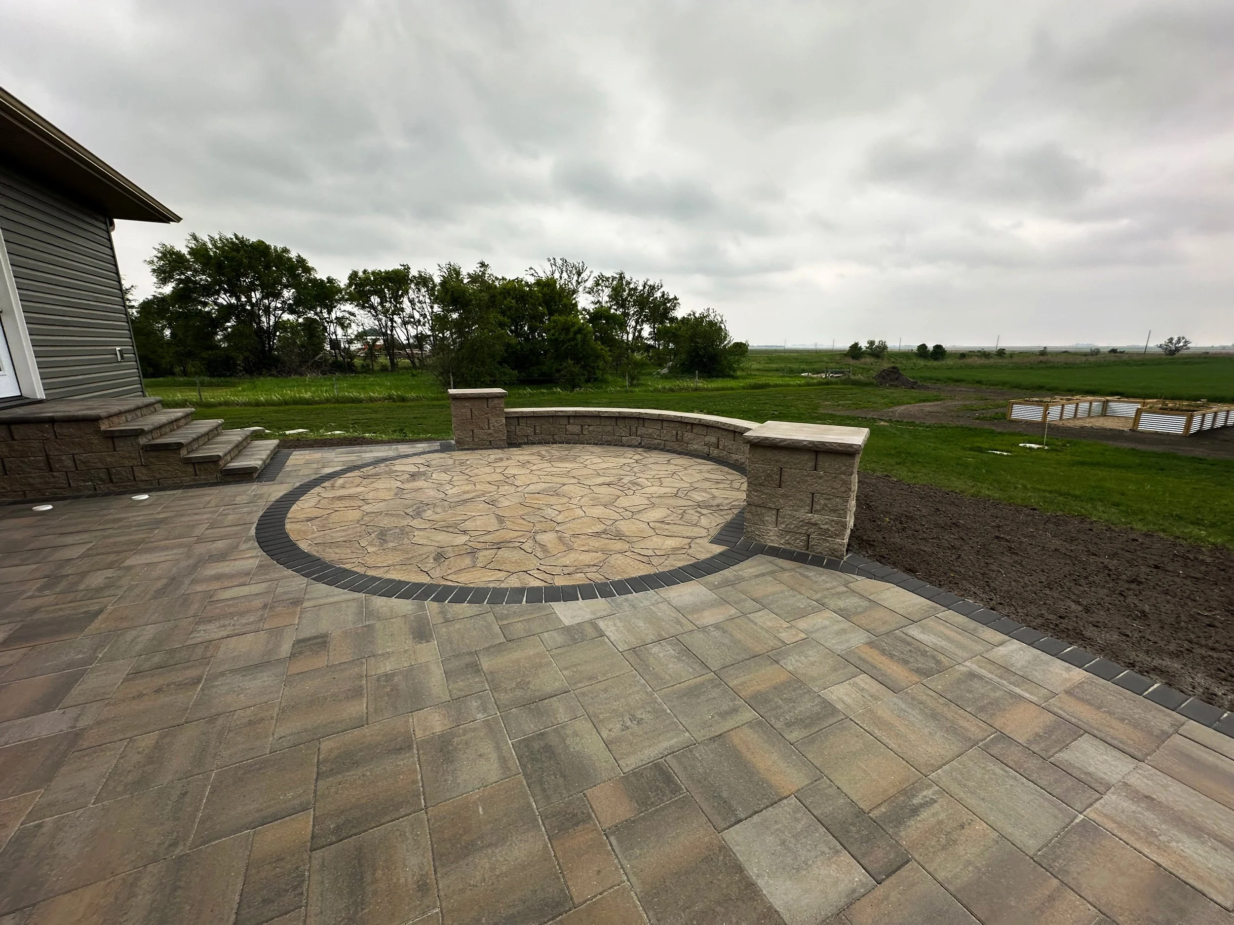 A newly constructed backyard patio with a circular stone design surrounded by pavers, adjacent to a house with steps, overlooking open green fields and cloudy sky.