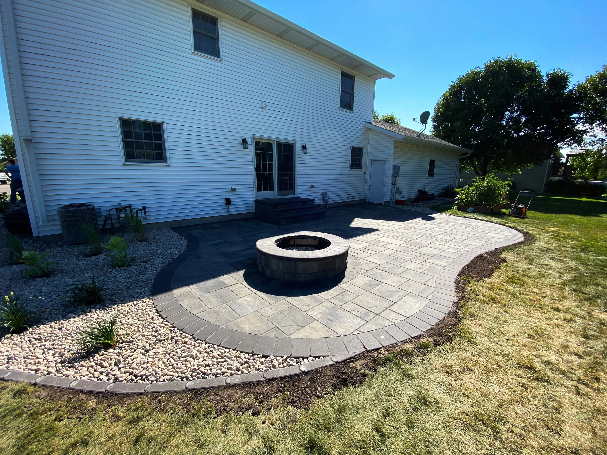 Backyard patio with stone and brick pavers, a fire pit in the center, and landscaped garden beds with small plants and rocks, adjacent to a white house with sliding glass door and multiple windows.