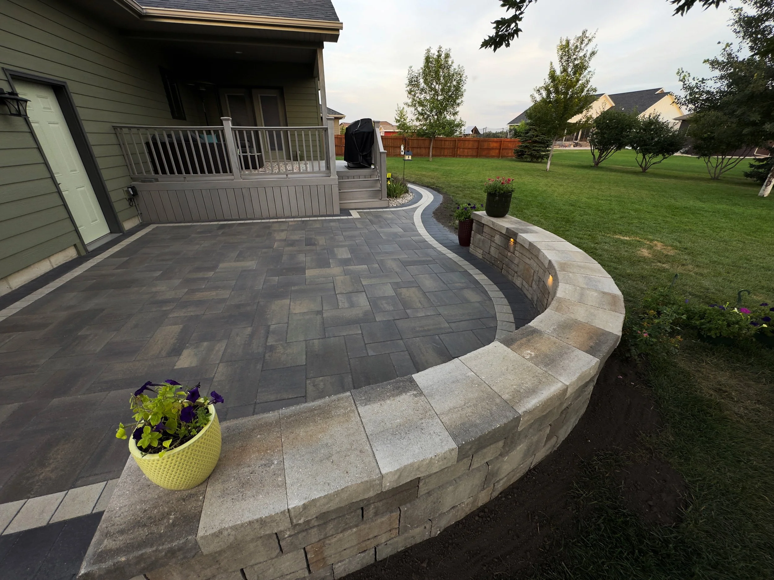 Backyard patio with paver stones, curved stone retaining wall, potted plants, grass lawn, trees, and a wooden fence in the background.