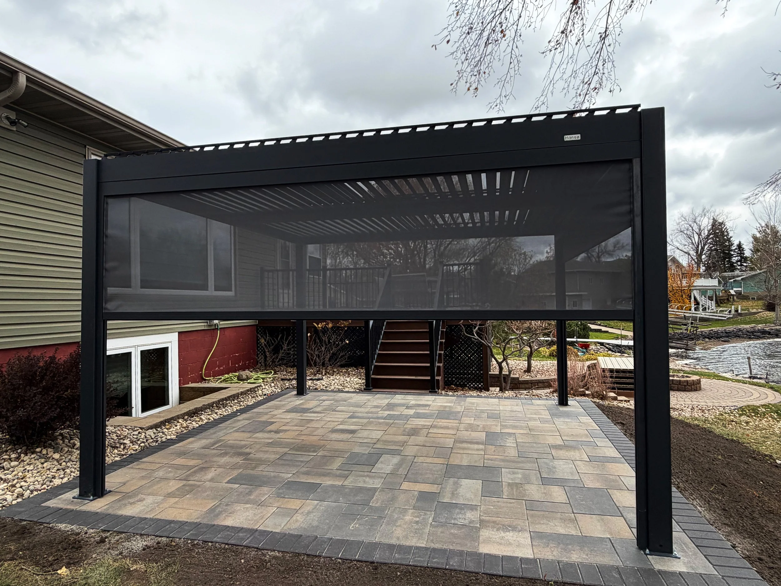 Patio area with a black metal carport with a roof structure, surrounding a paved stone surface, next to a house with siding, toward a lake or pond with a curved seating area and trees in the background.