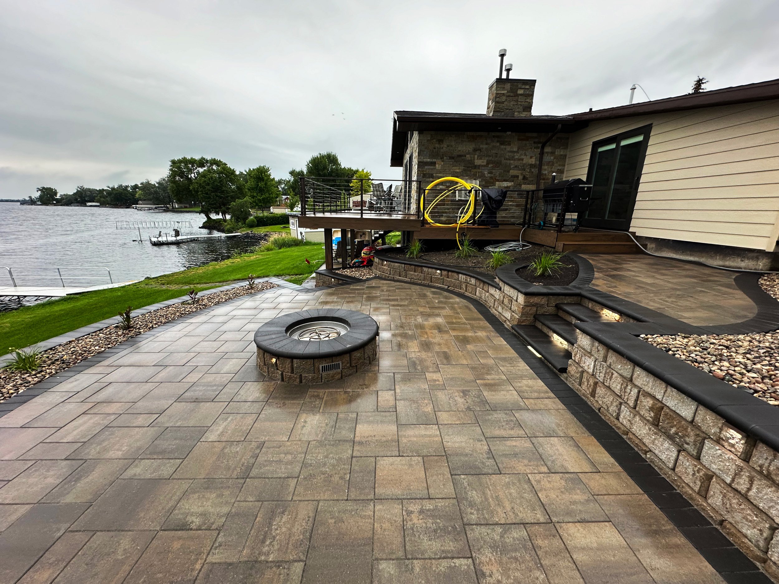 L-shaped backyard patio with stone pavers, a fire pit, landscaped flower beds with small plants, and a deck overlooking a lake with a dock, surrounded by grass and trees.