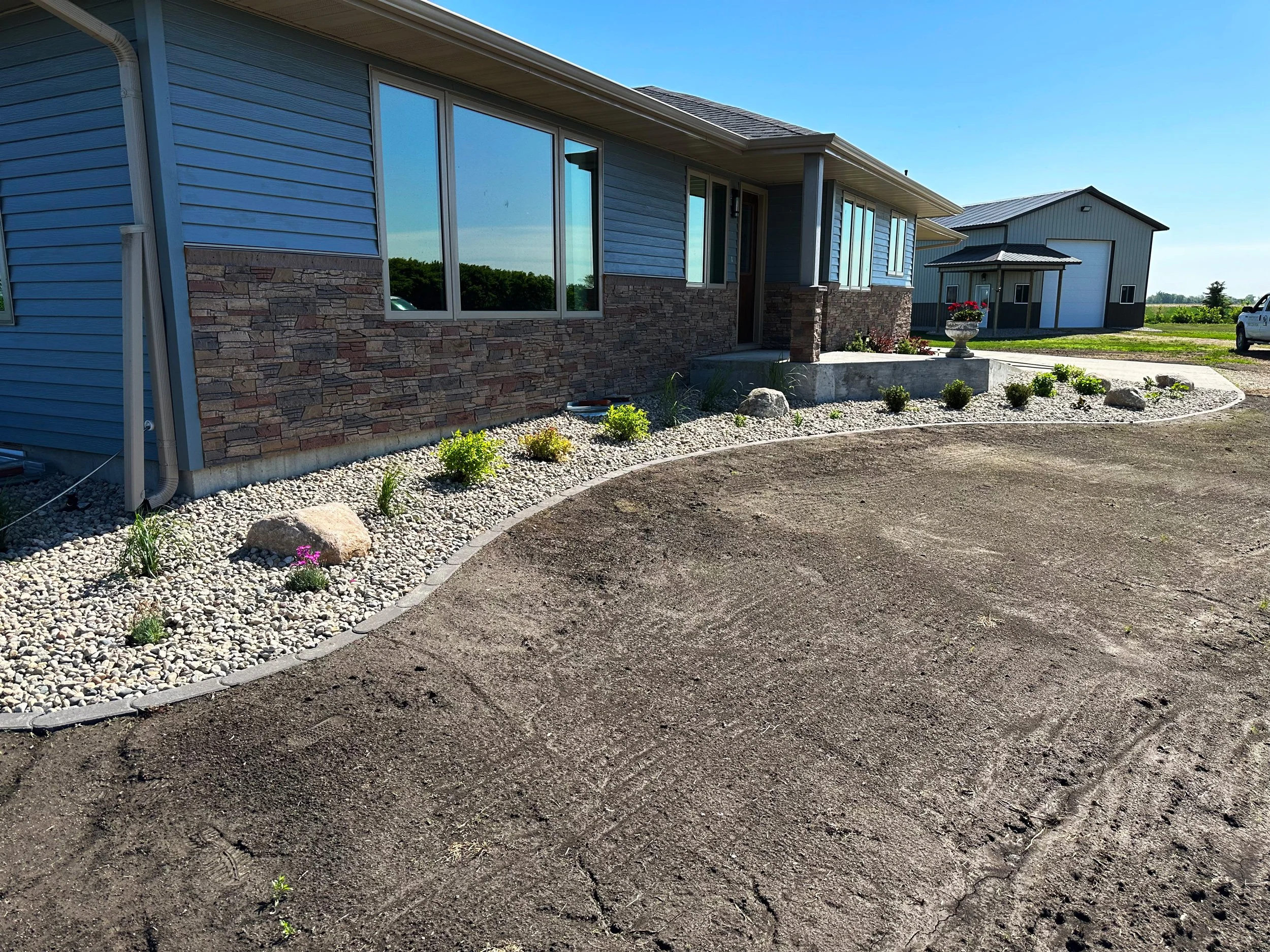 View of a house with blue siding, stone accents, and large windows. The front yard has a curved gravel pathway with small plants and rocks, a concrete step, and a decorative urn. There is a driveway with tire tracks and a neighboring metal building i