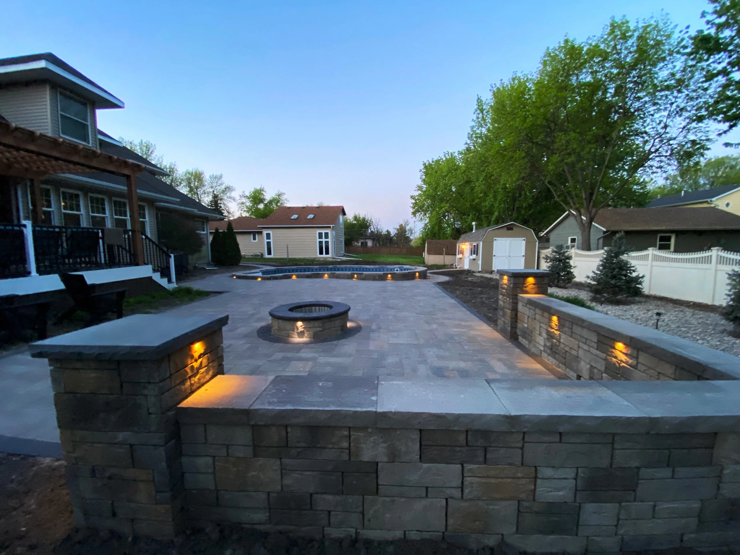 A backyard patio with stone walls, outdoor lighting, a fire pit, and a swimming pool in the background, surrounded by trees and neighboring houses.