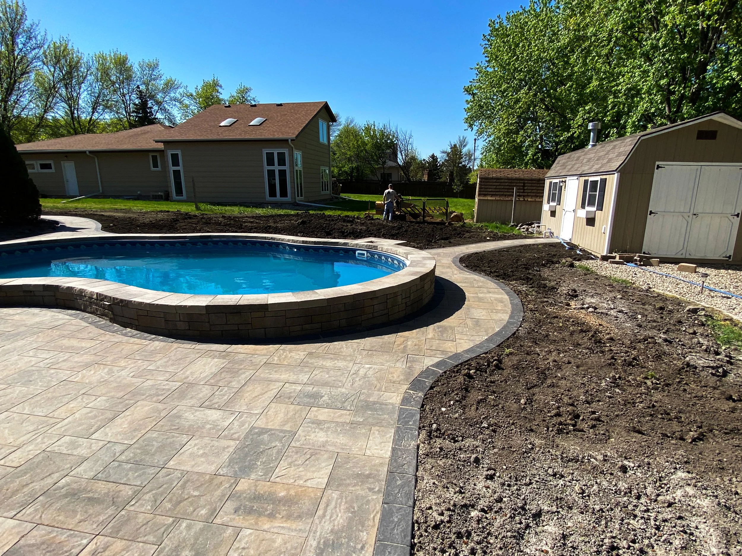 An outdoor backyard scene with a new in-ground swimming pool with a stone brick border, surrounded by a stamped concrete patio. There are patches of dirt and a garden bed, and in the background, a house, a shed, and trees under a clear blue sky.