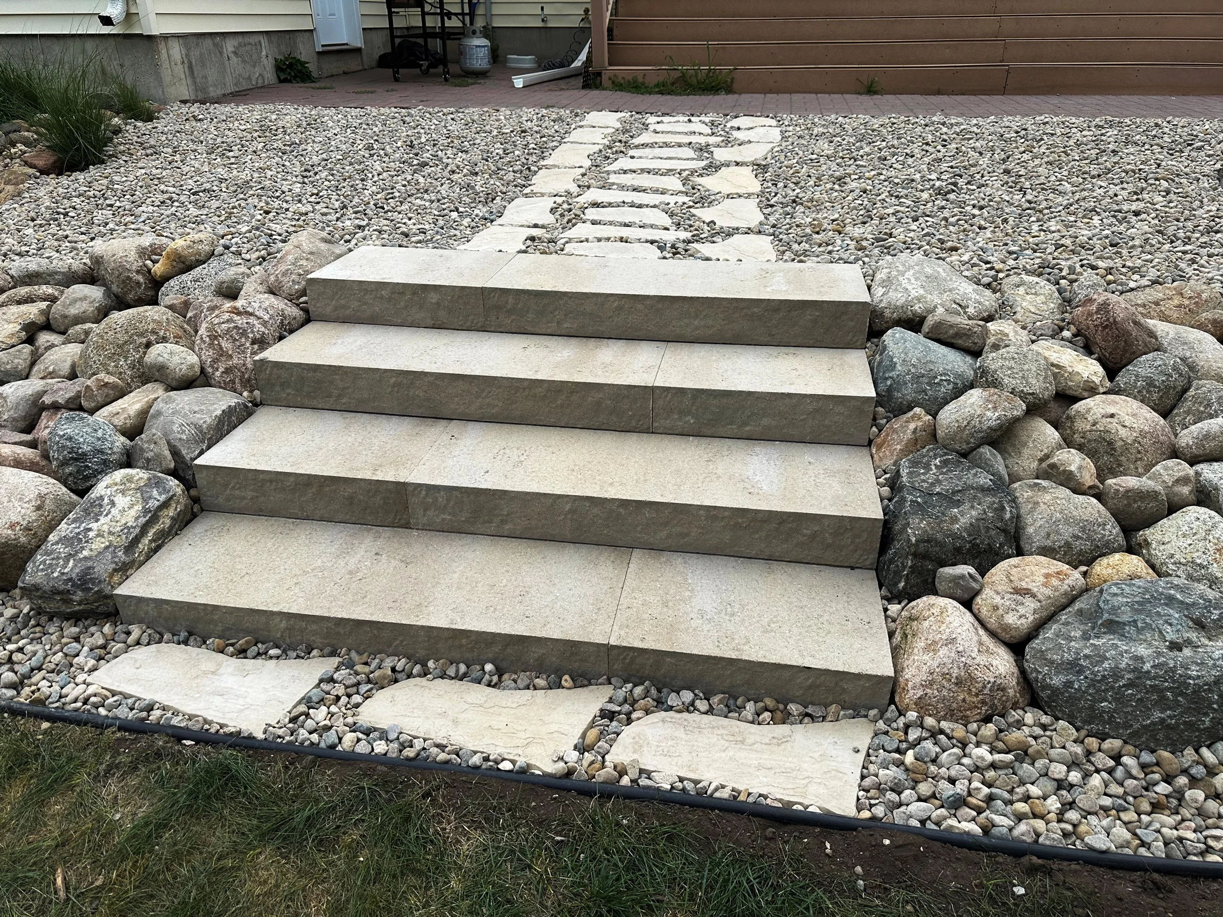 Concrete steps leading up to a walkway with large rocks on sides, surrounded by gravel and decorative stones in a yard.