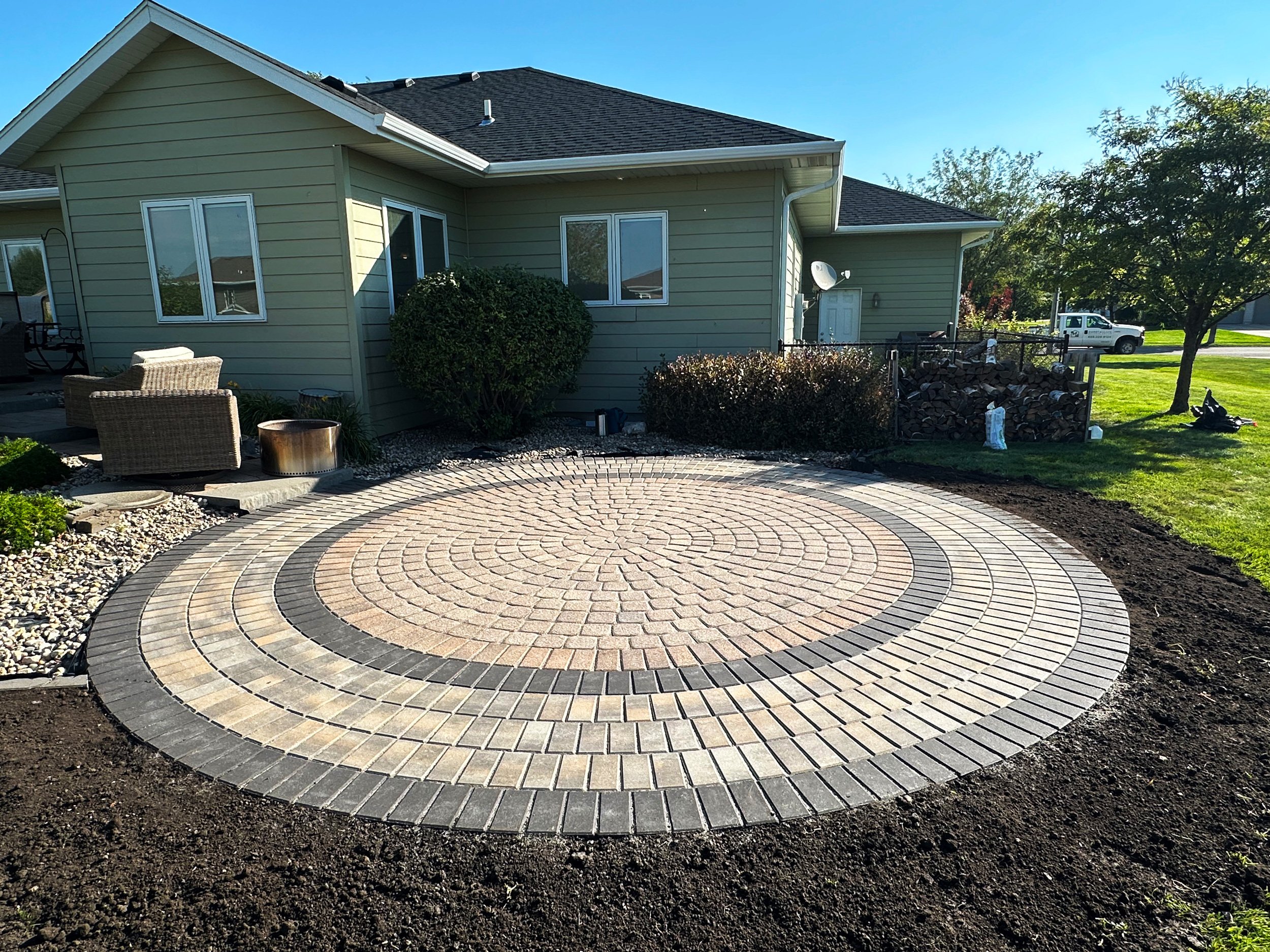 Circular brick patio in the backyard of a house with green siding, outdoor seating, and a woodpile, under a blue sky.