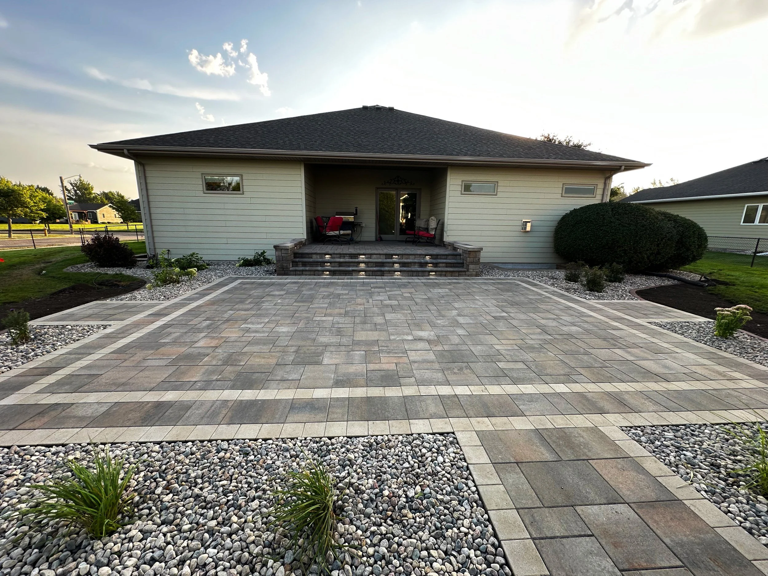 Backyard with newly paved stone patio, steps leading to a house with a porch, outdoor seating, and landscaped garden beds with rocks and small plants.