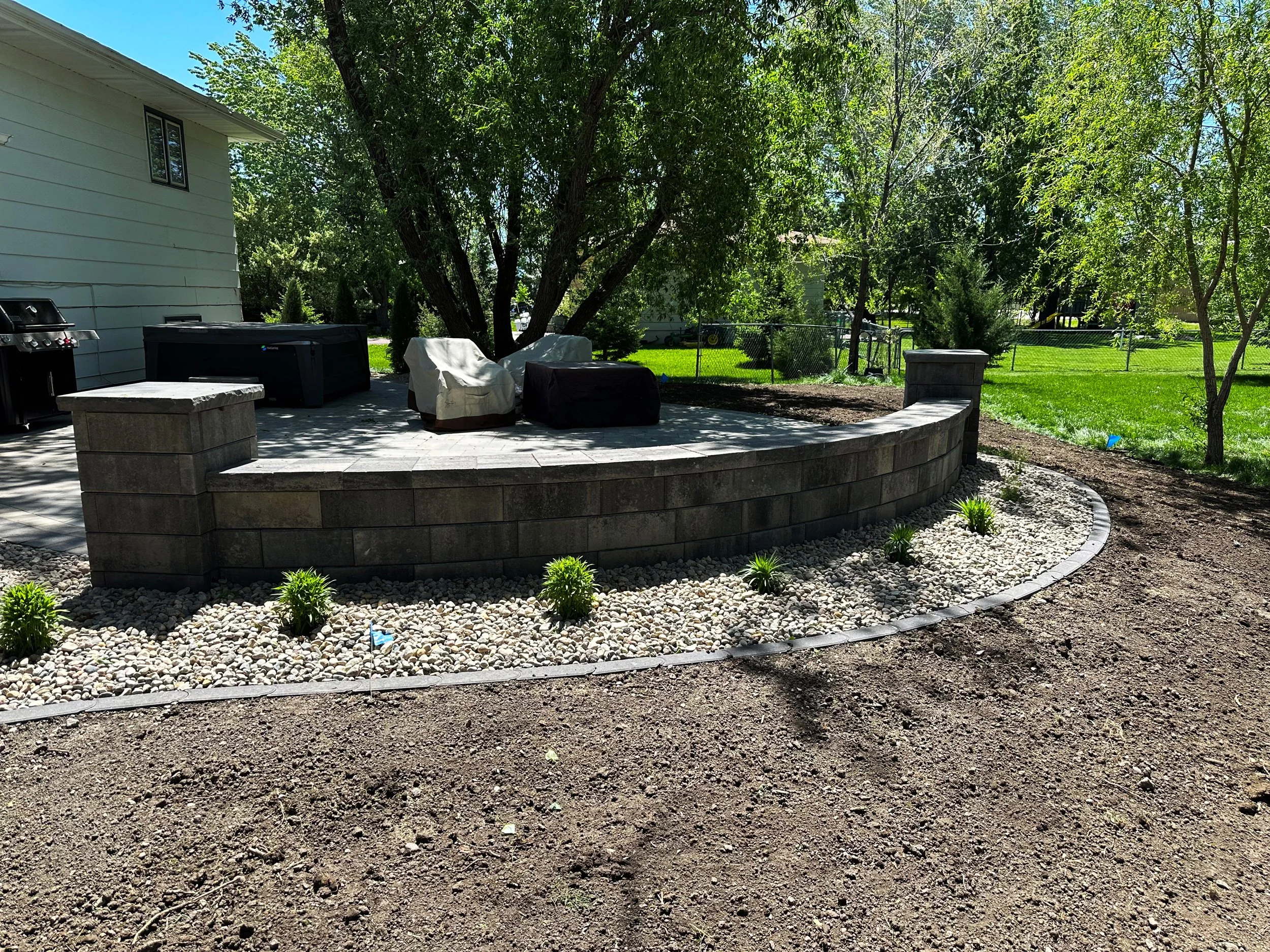 Backyard patio with stone retaining wall, covered outdoor furniture, and barbecue grill, surrounded by trees and grass.