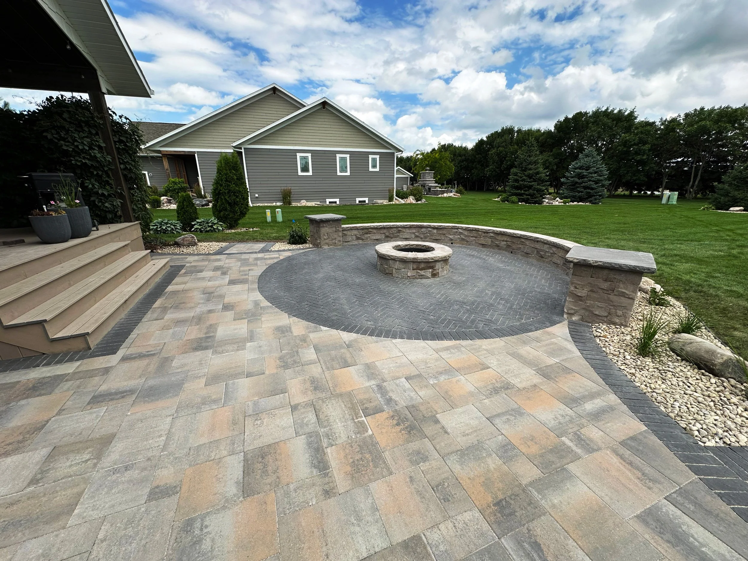 Backyard with paved patio, stone fire pit, green lawn, trees, and a house with gray siding under a partly cloudy sky.