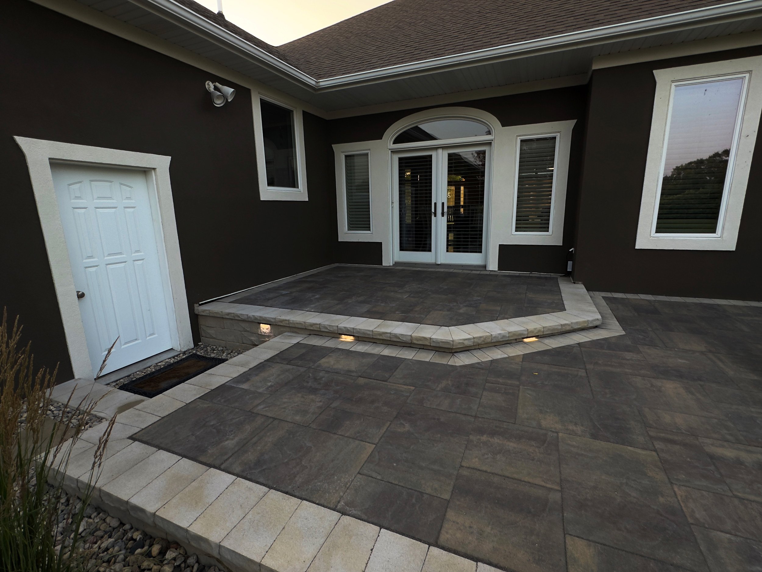 Backyard patio with a raised stone deck, surrounding tiled flooring, and sliding glass doors leading into the house. There are two windows and a small white door on the dark-colored exterior wall.