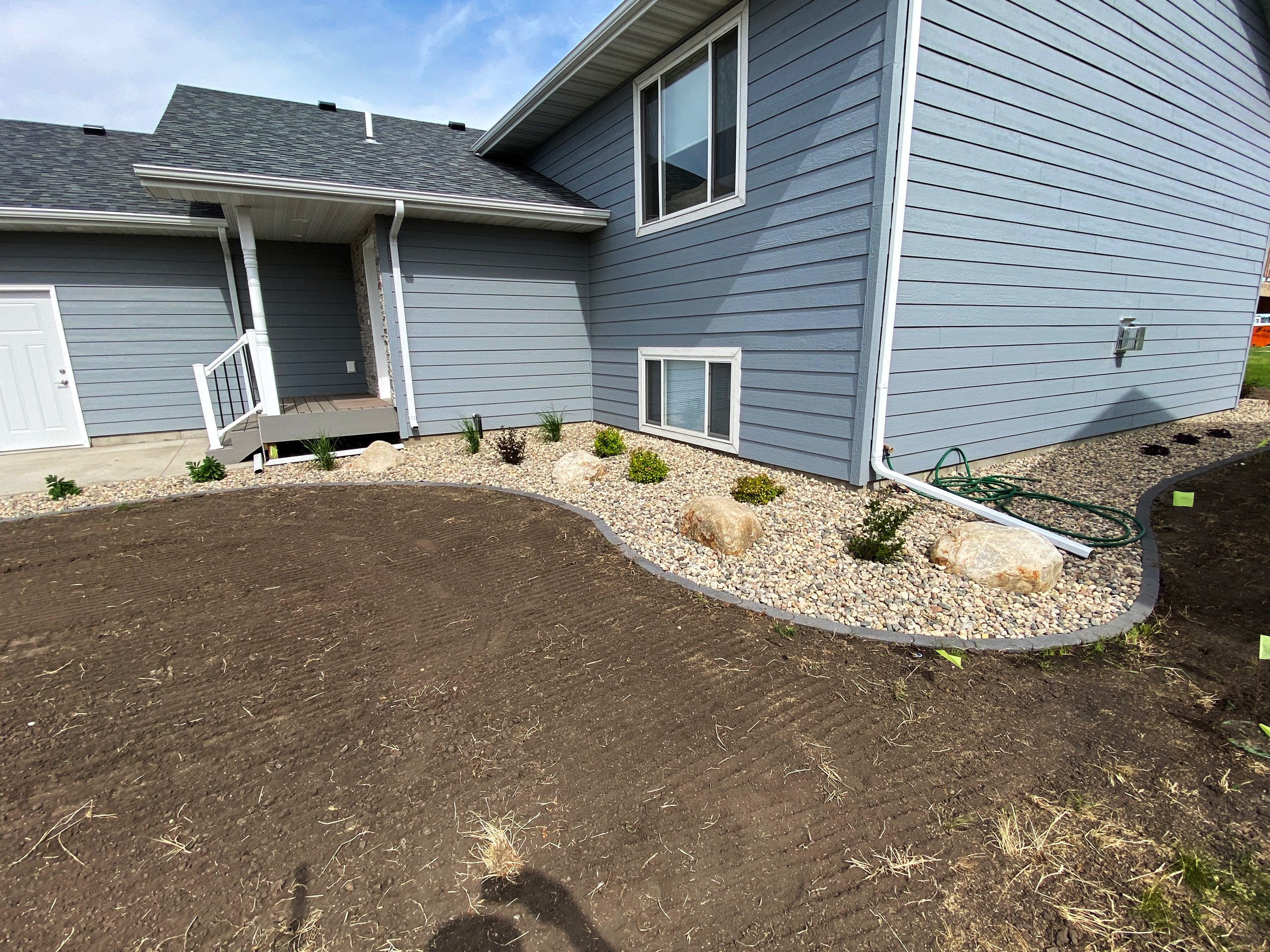 Backyard landscaping with a freshly planted flower bed with rocks, small shrubs, and soil, next to a light blue house with white trim and several small basement windows.
