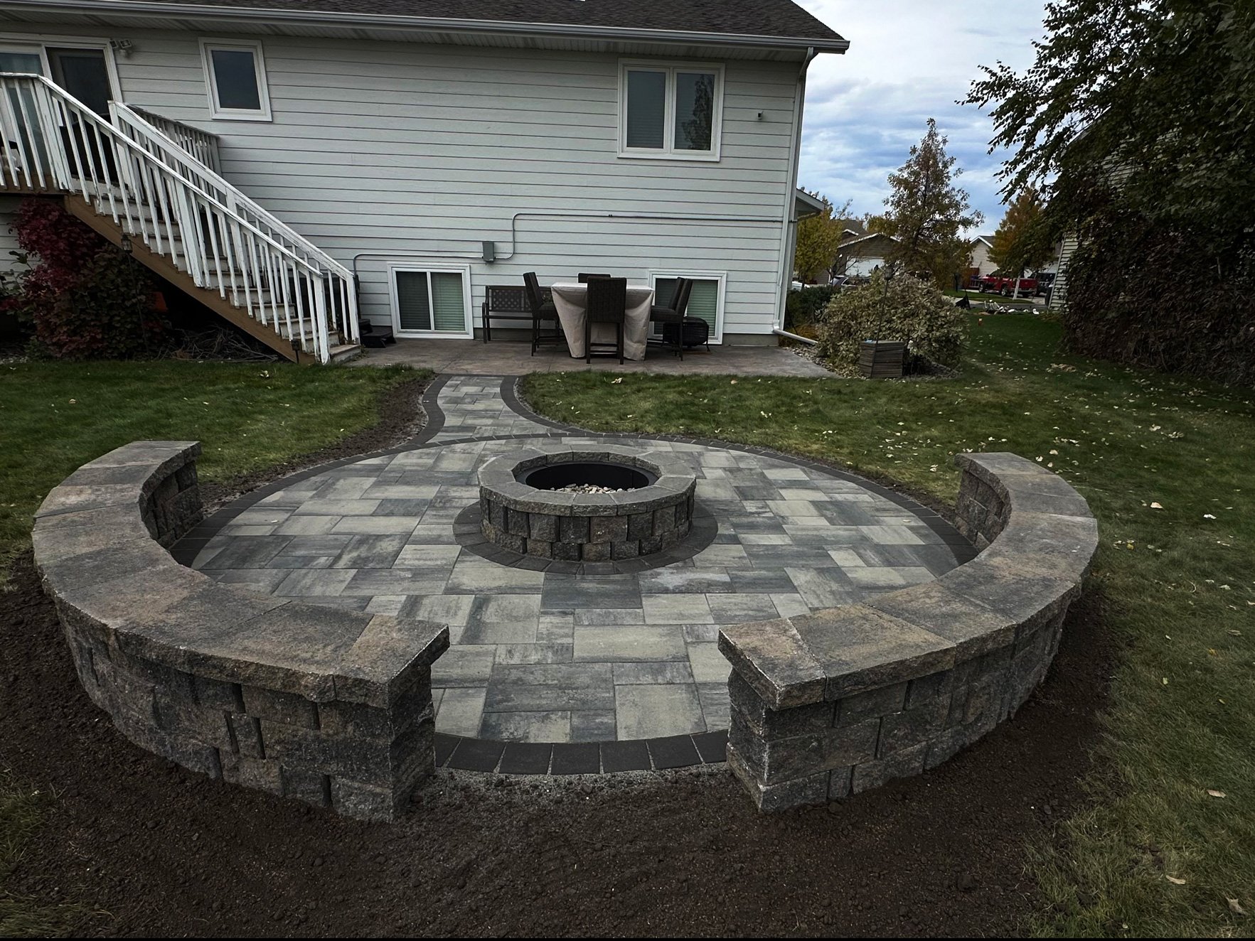 Backyard patio with a stone fire pit in the center, surrounded by curved stone seating, and a patterned stone walkway leading to a white house with a deck and outdoor furniture.