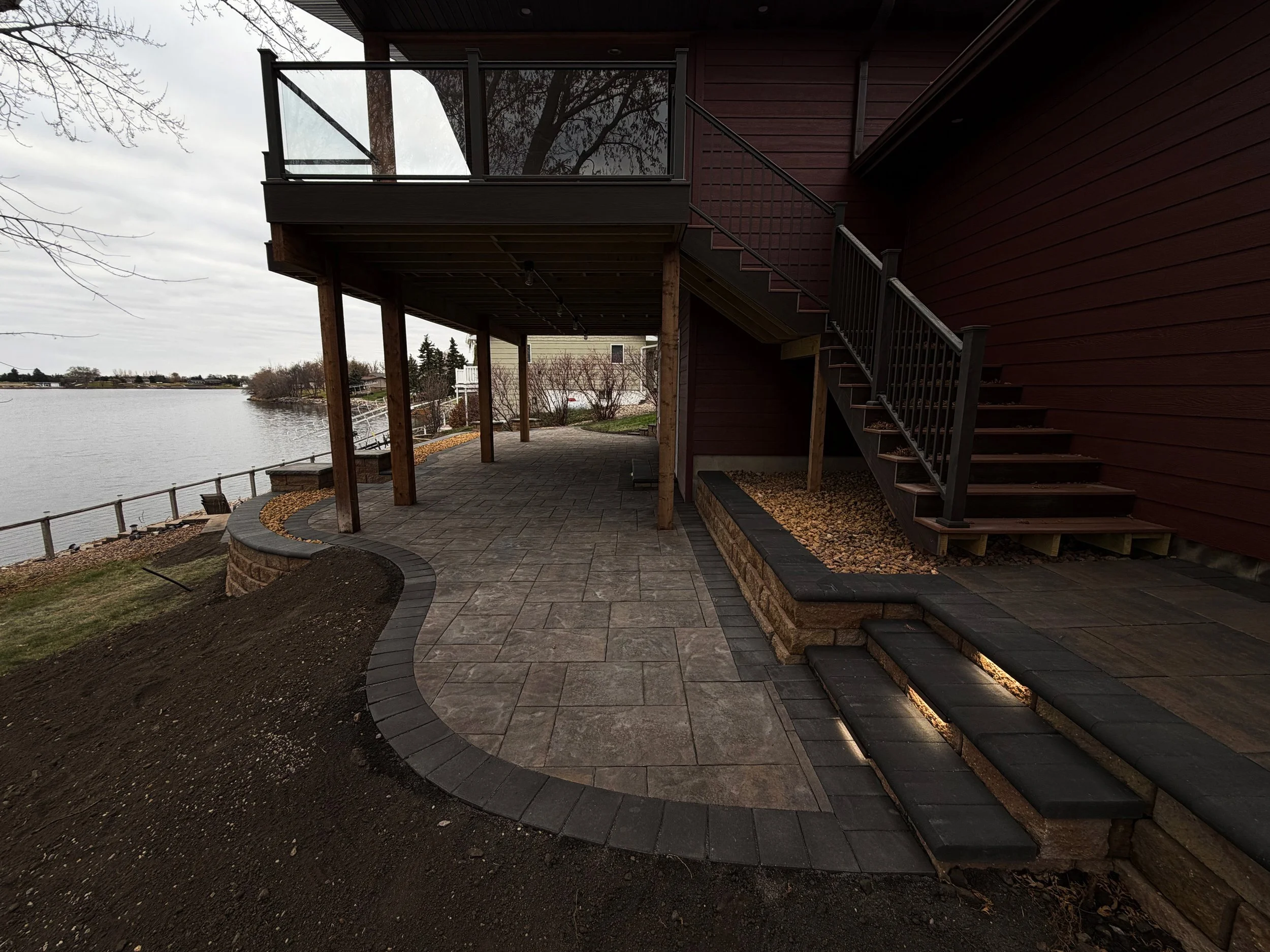 A backyard patio by a river with a raised deck, stairs, and stone pavers. The patio has a curved edging with some gardening beds, and a railing along the water's edge. The sky is overcast.