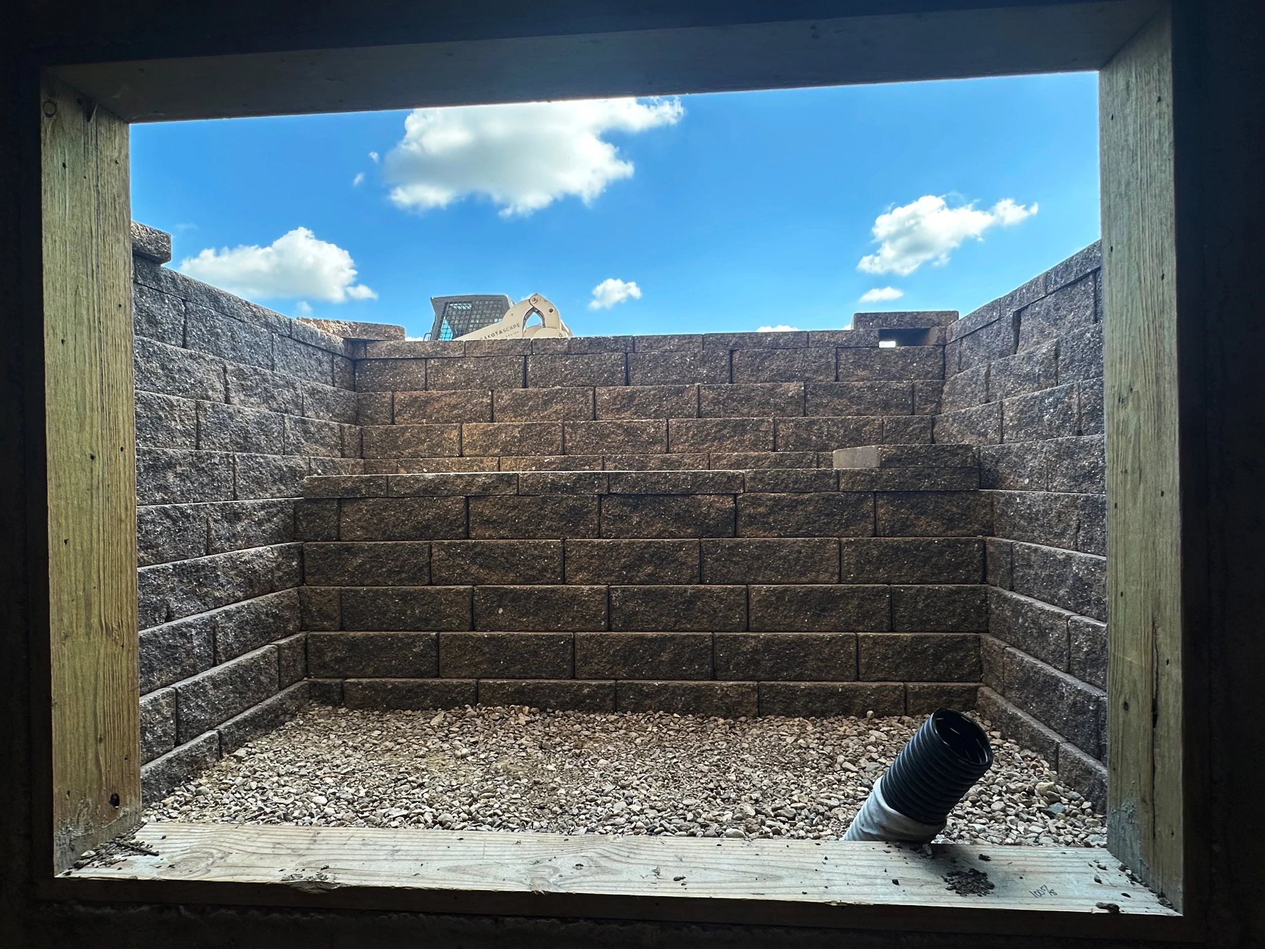 View of an outdoor construction site through a rectangular opening. There are brown brick walls, gravel on the ground, and tools like a black pipe and a plastic container visible. Blue sky with clouds is seen above.