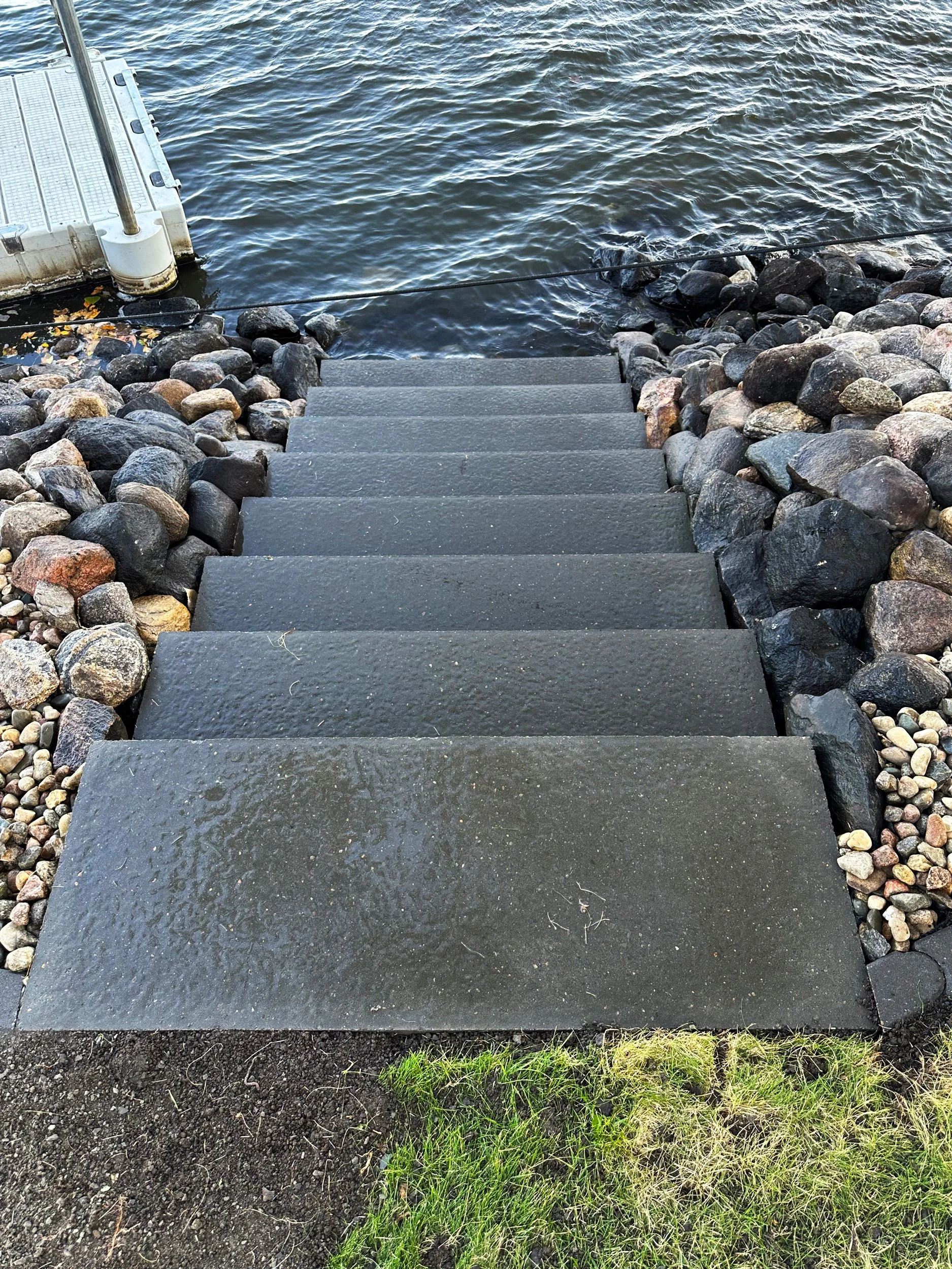 Concrete steps leading down to a body of water, likely a lake or river, surrounded by rocks and pebbles, with a grassy area at the bottom of the stairs.