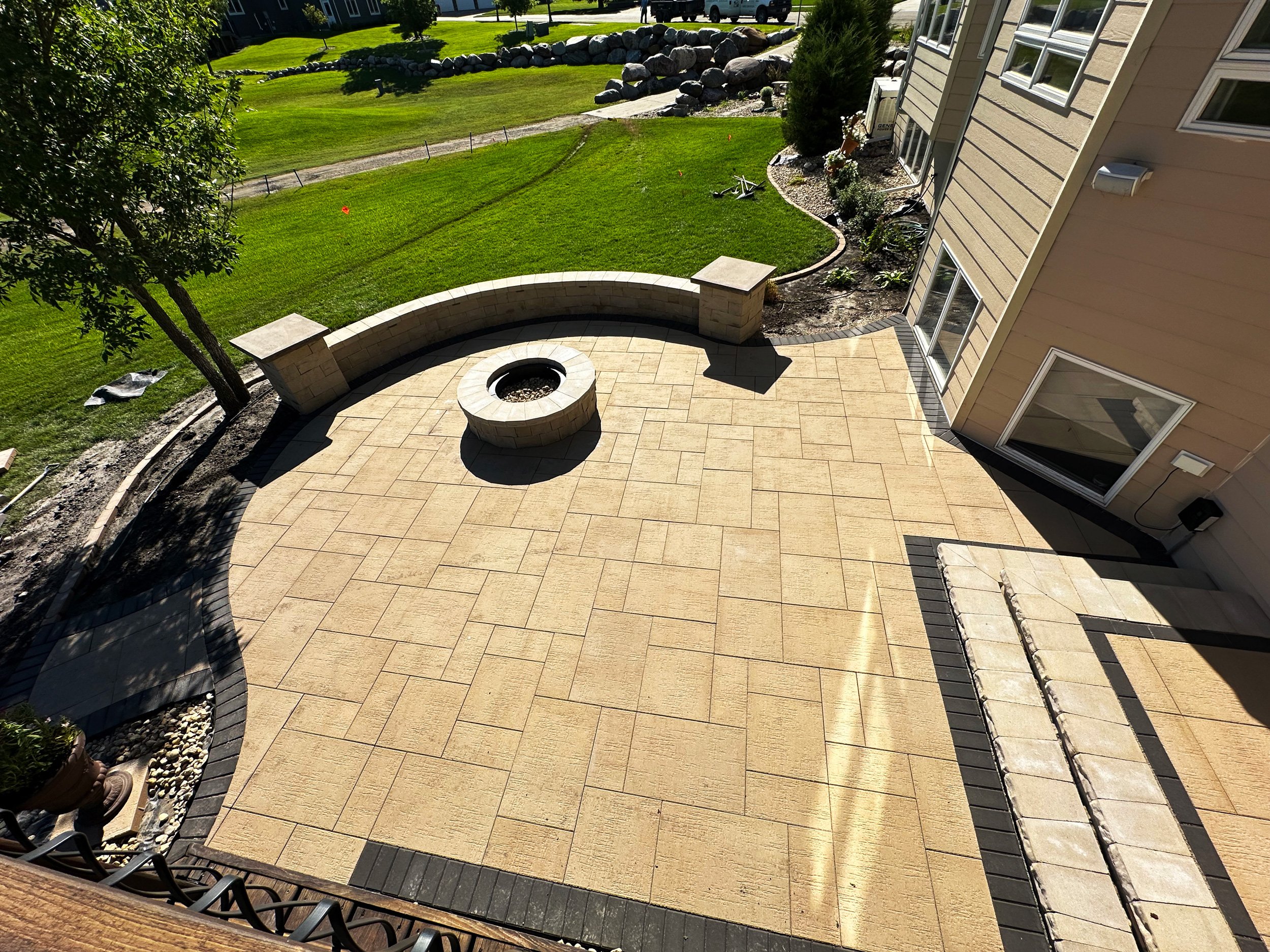 A backyard with a stone patio, a fire pit, a curved stone bench, and a grassy area with trees and rocks, viewed from an elevated perspective.