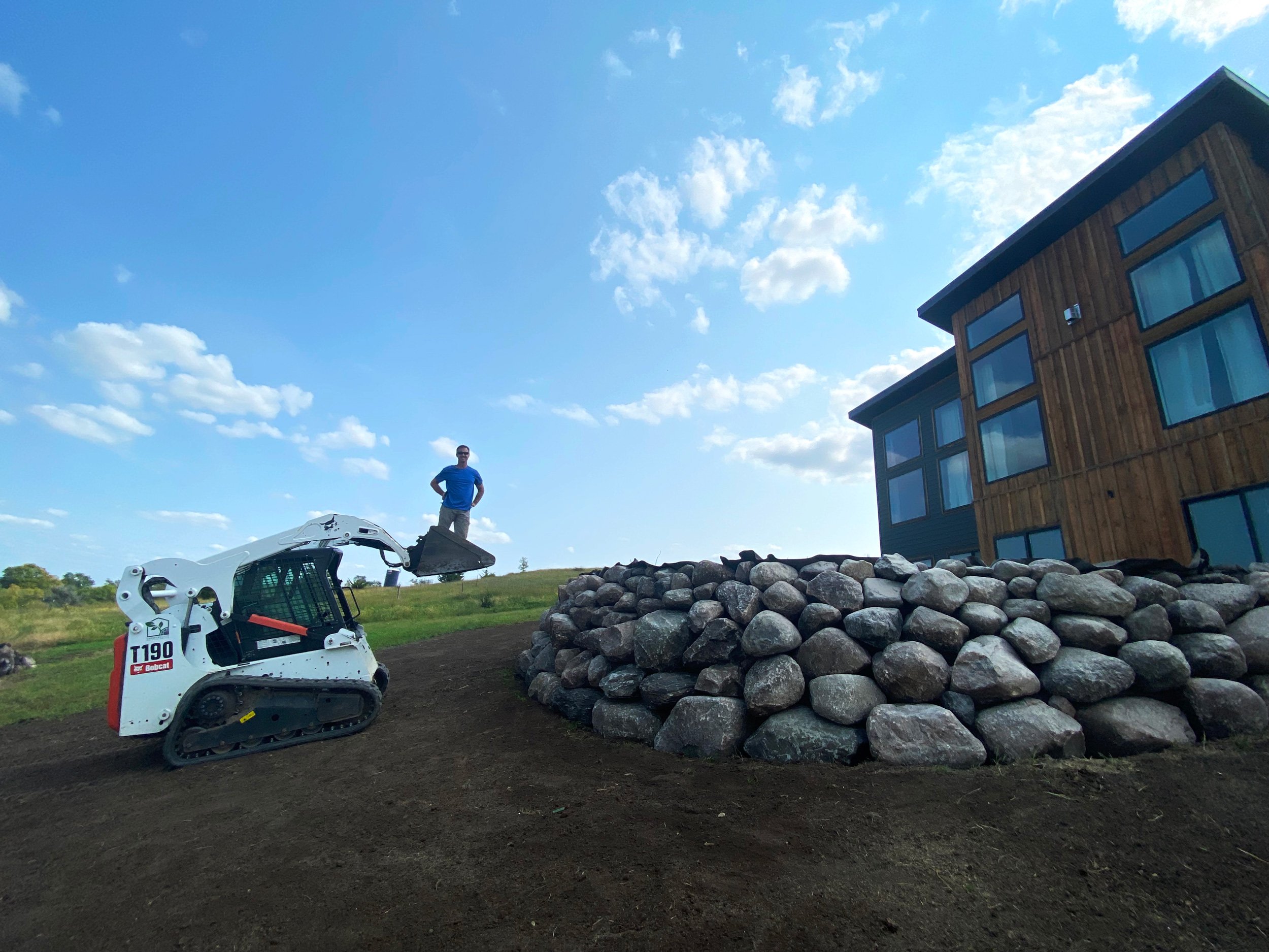 A man standing on a grassy hill next to a small white tracked Bobcat T190 skid-steer loader with a bucket, next to a large pile of rocks and a modern wooden house with large windows under a blue sky with scattered clouds.