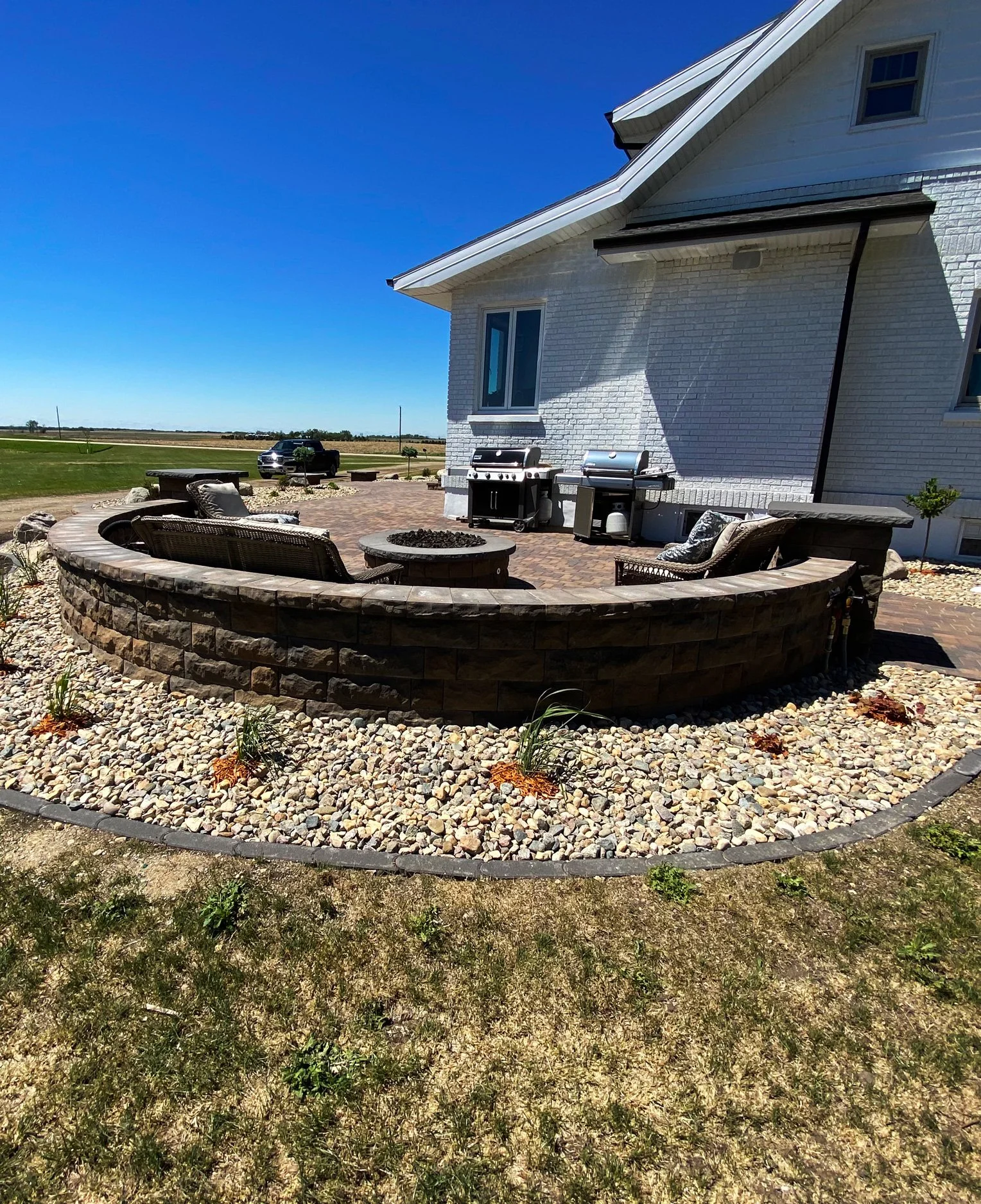 Outdoor patio area with a circular stone fire pit, wicker chairs with cushions, and a brick house in the background with grills and a large window.