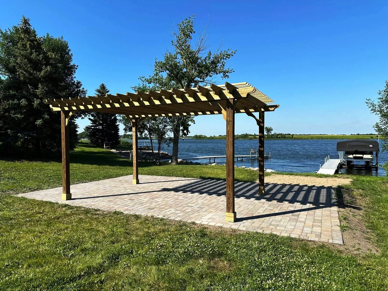 A wooden pergola with a brick patio, overlooking a lake with a boat dock and boat, surrounded by trees and green grass under a clear blue sky.