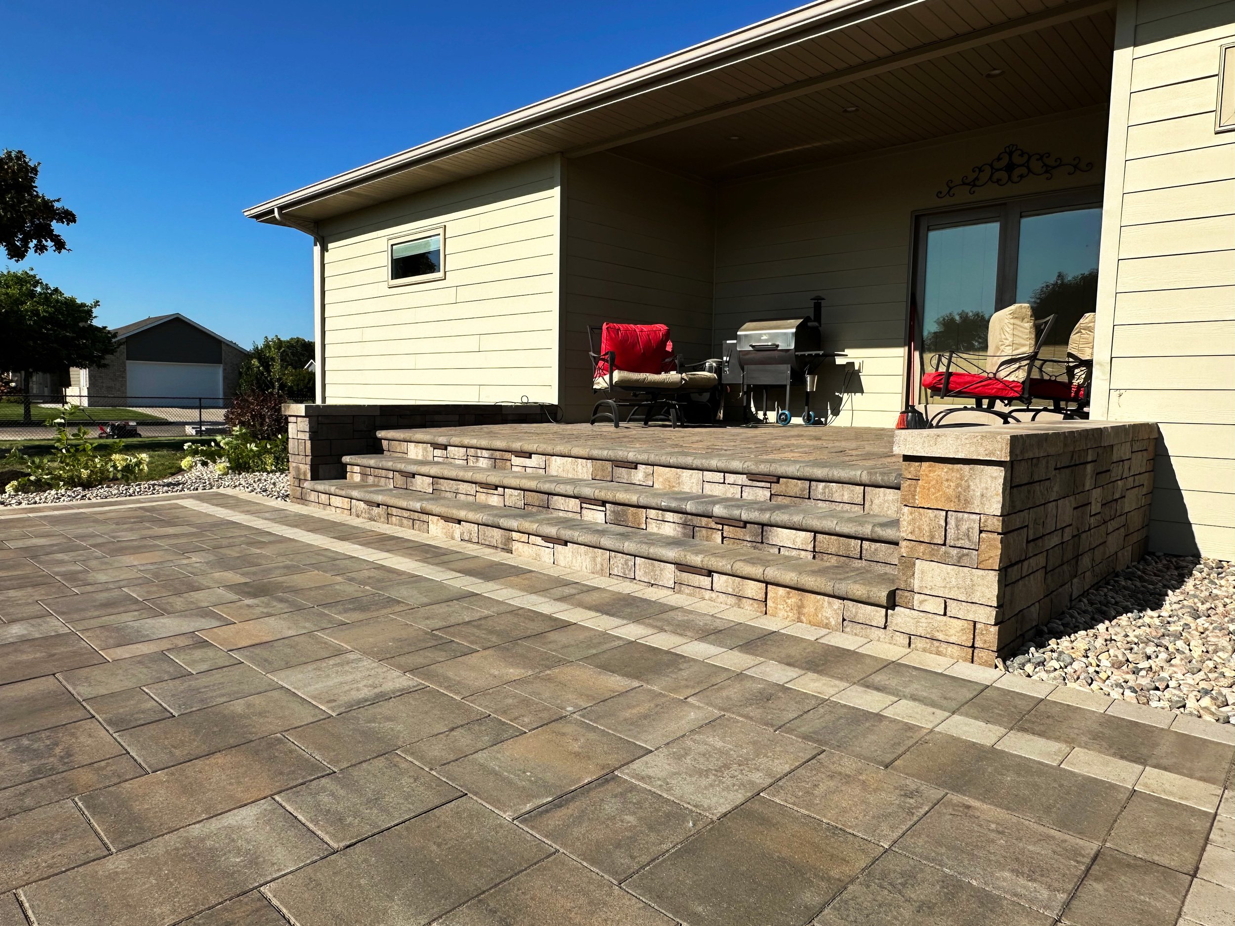 Back porch of a house with stairs leading to a paved patio, outdoor chairs, a barbecue grill, and garden area with plants and rocks.