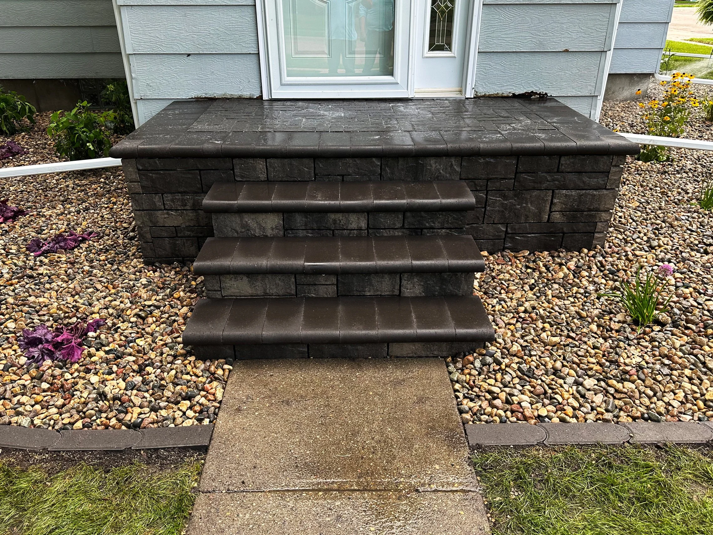 Front porch with four dark brown steps leading to a white front door with a decorative glass window, gray siding on the house, and a flower bed with colorful flowers and small rocks on either side of the steps.