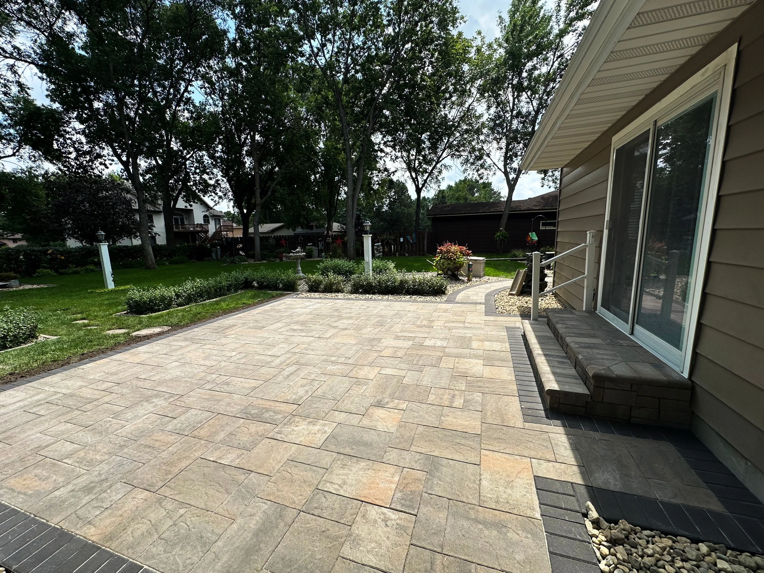 A backyard patio with beige pavers, adjacent to a house with tan siding and a glass sliding door. The yard has green grass, trees, and decorative plants, with a garden bed and pathway in the background.