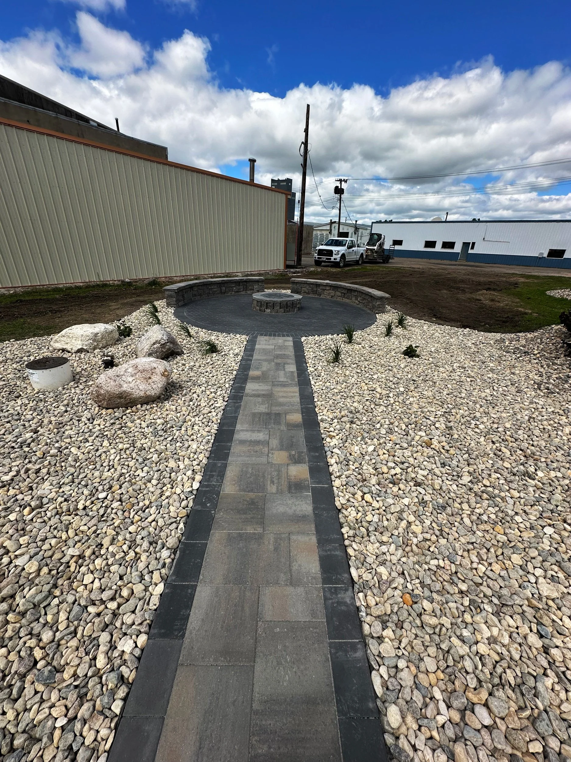 A landscaped outdoor area with a paved walkway leading to a circular seating area with stone benches, rocks, and small plants, under a cloudy sky.