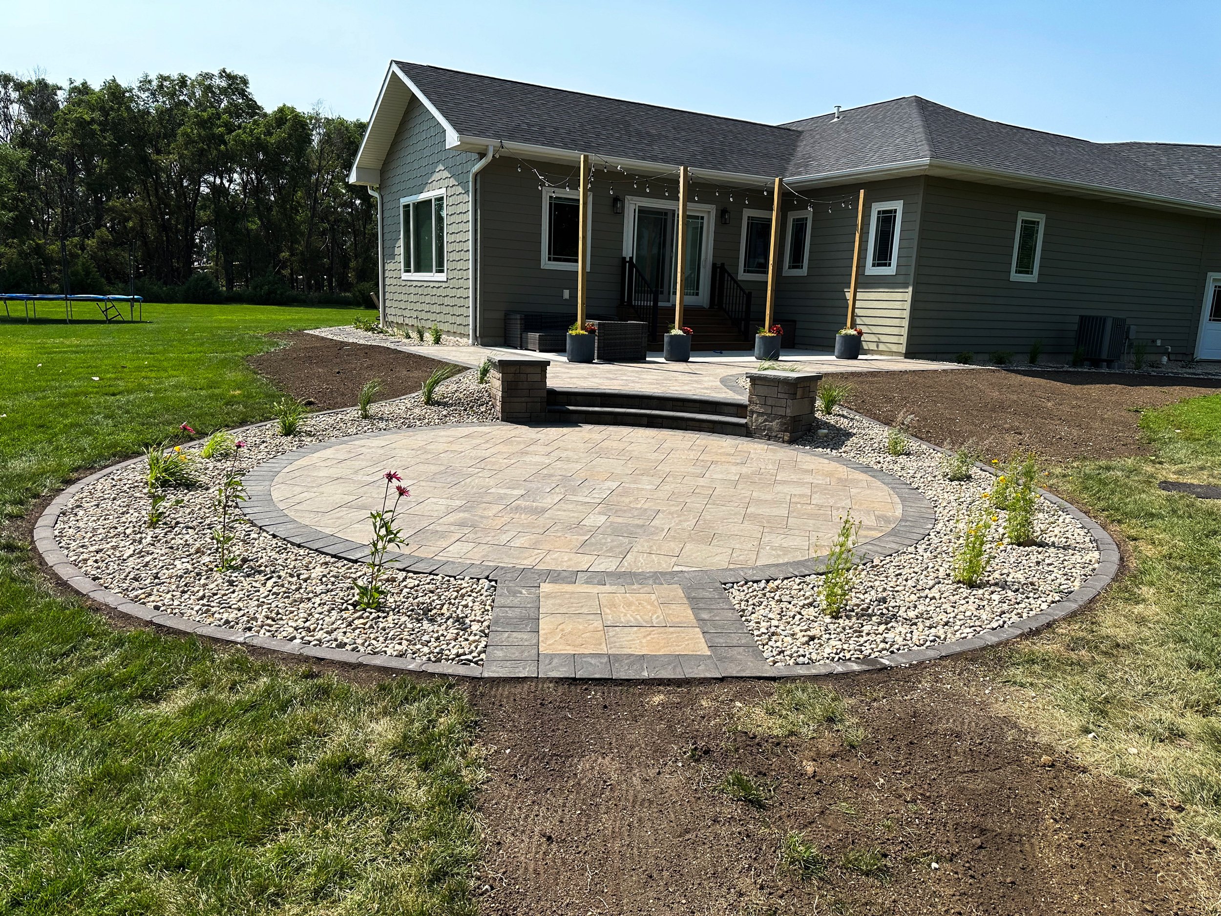 Backyard patio with circular stone paver design, small plants, steps leading to house, and a deck with string lights, surrounded by lawns and a wooded area.