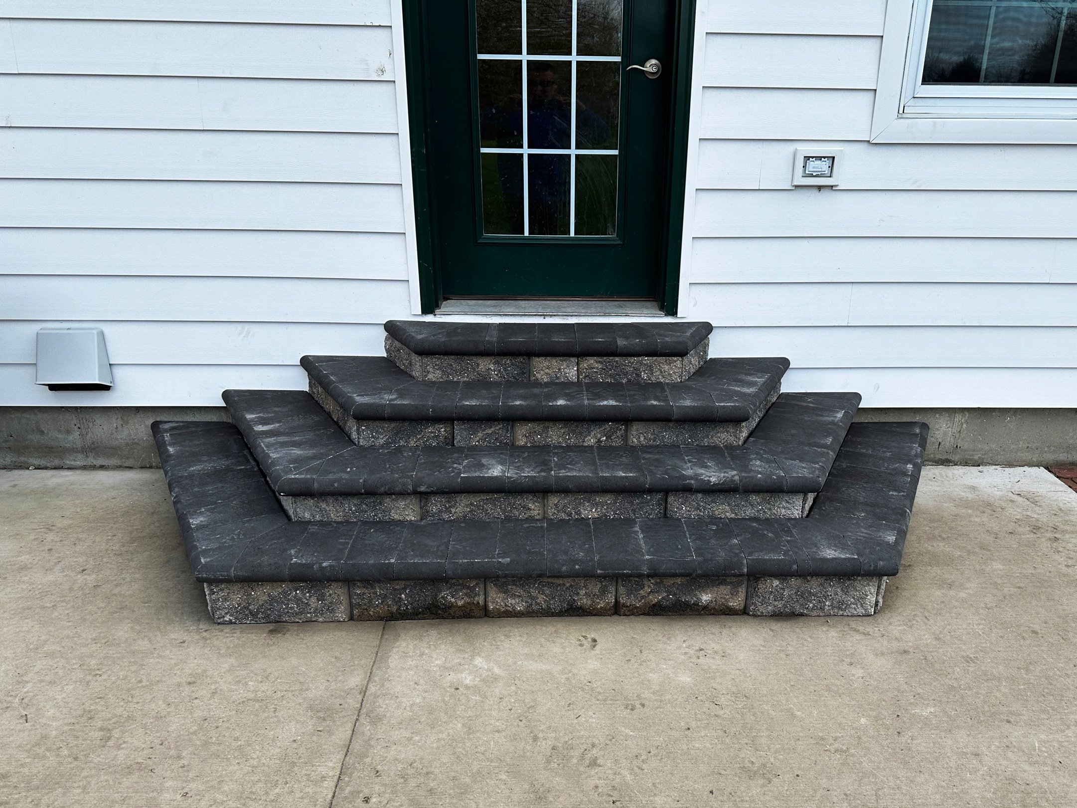 Stone steps leading up to a green door on a white house with horizontal siding, concrete sidewalk in front.