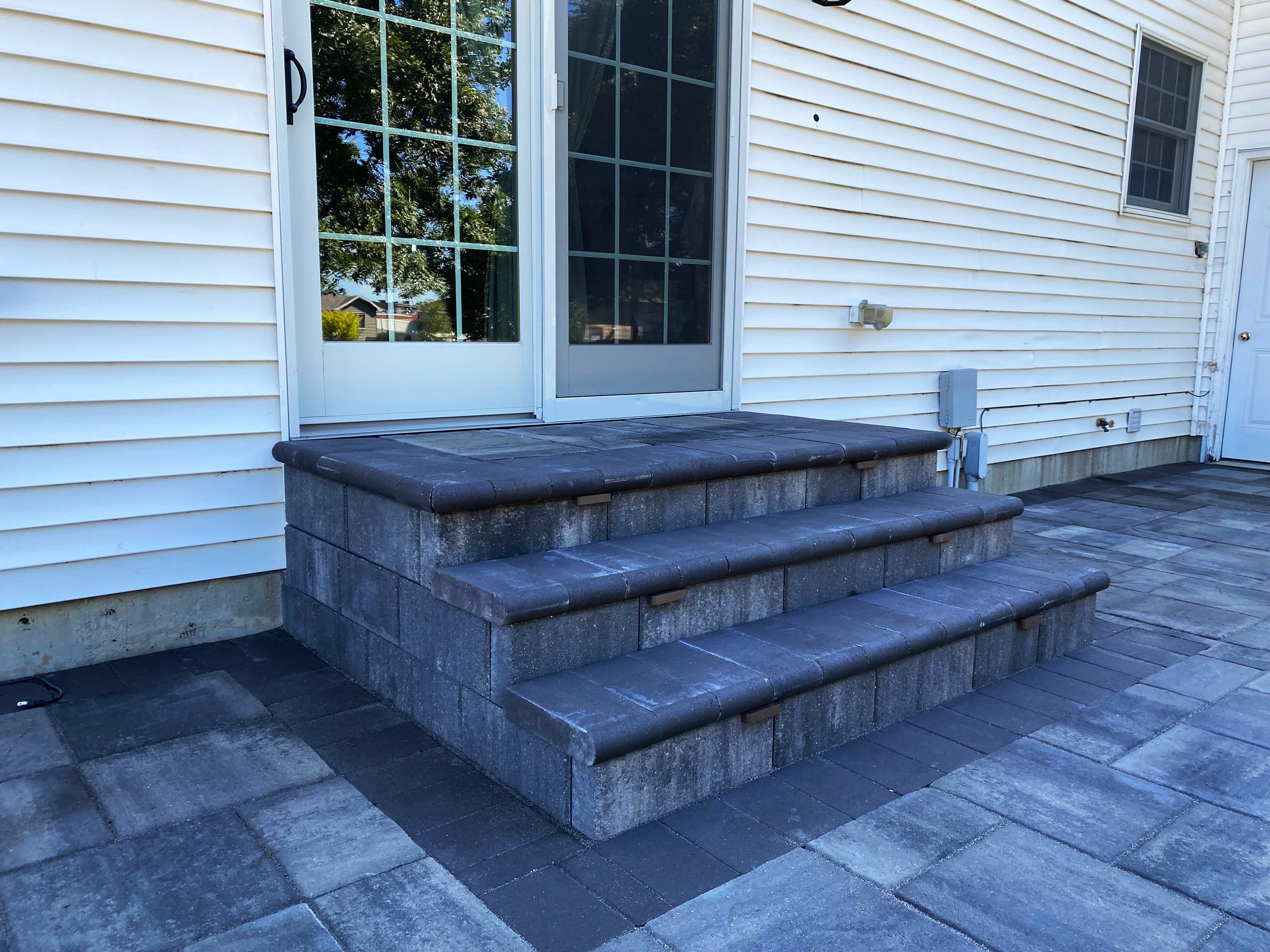 Backyard patio with dark gray stone steps leading up to a glass door, beige house siding, and a paved area with gray and black stones.