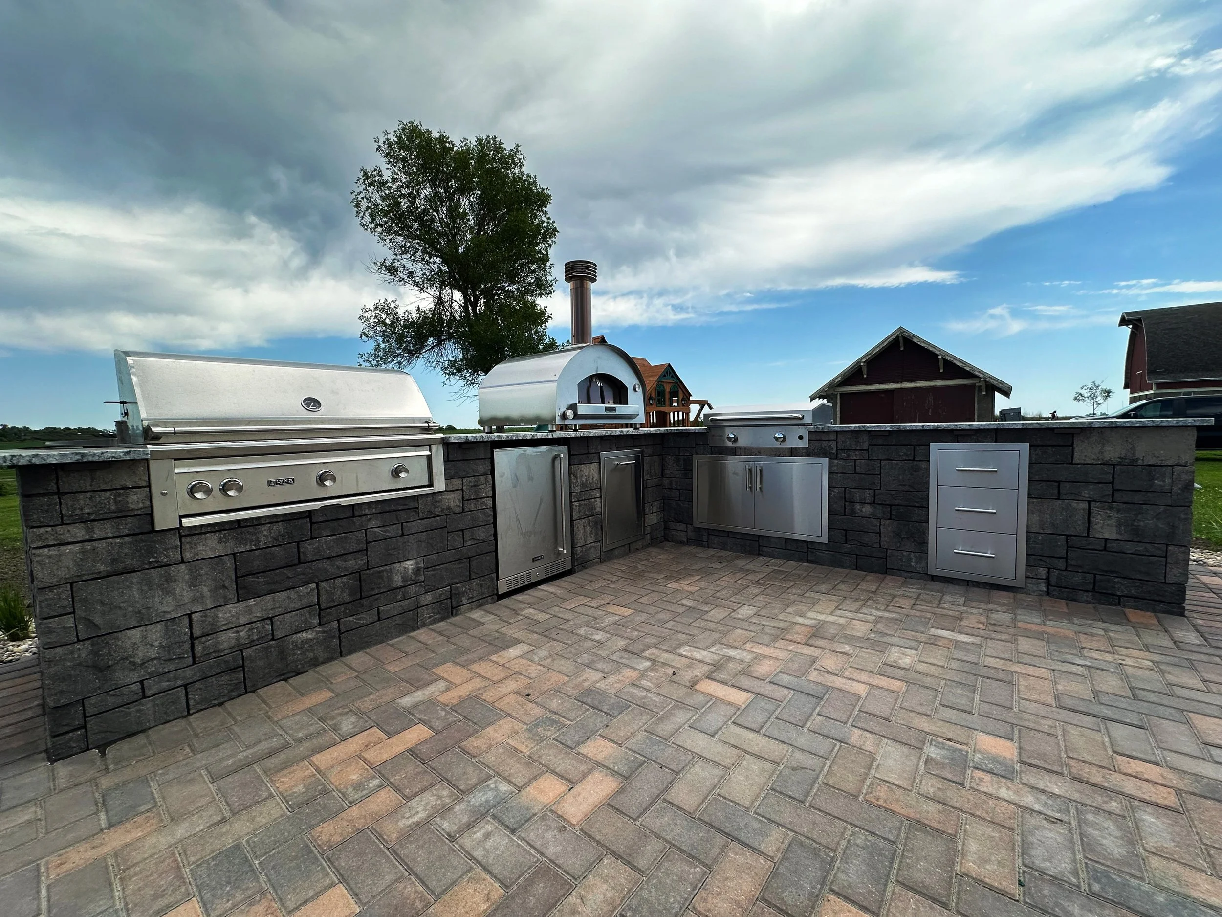 Outdoor kitchen with stainless steel appliances, a grill, a pizza oven, and storage cabinets, set on brick pavers with a cloudy sky, trees, and buildings in the background.
