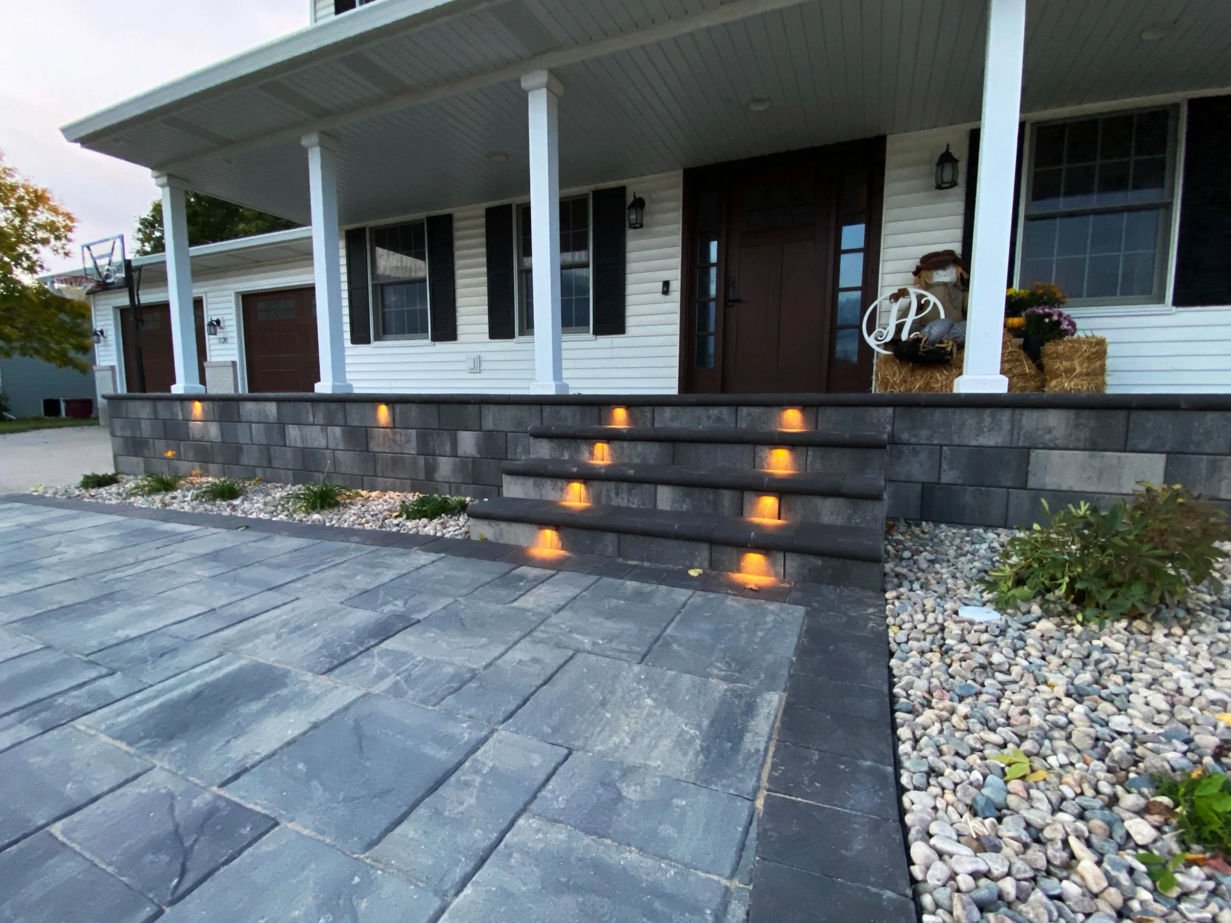Front porch of a house with steps and stone walkway, landscape lighting, potted plants, and decorative hay bales with flowers.