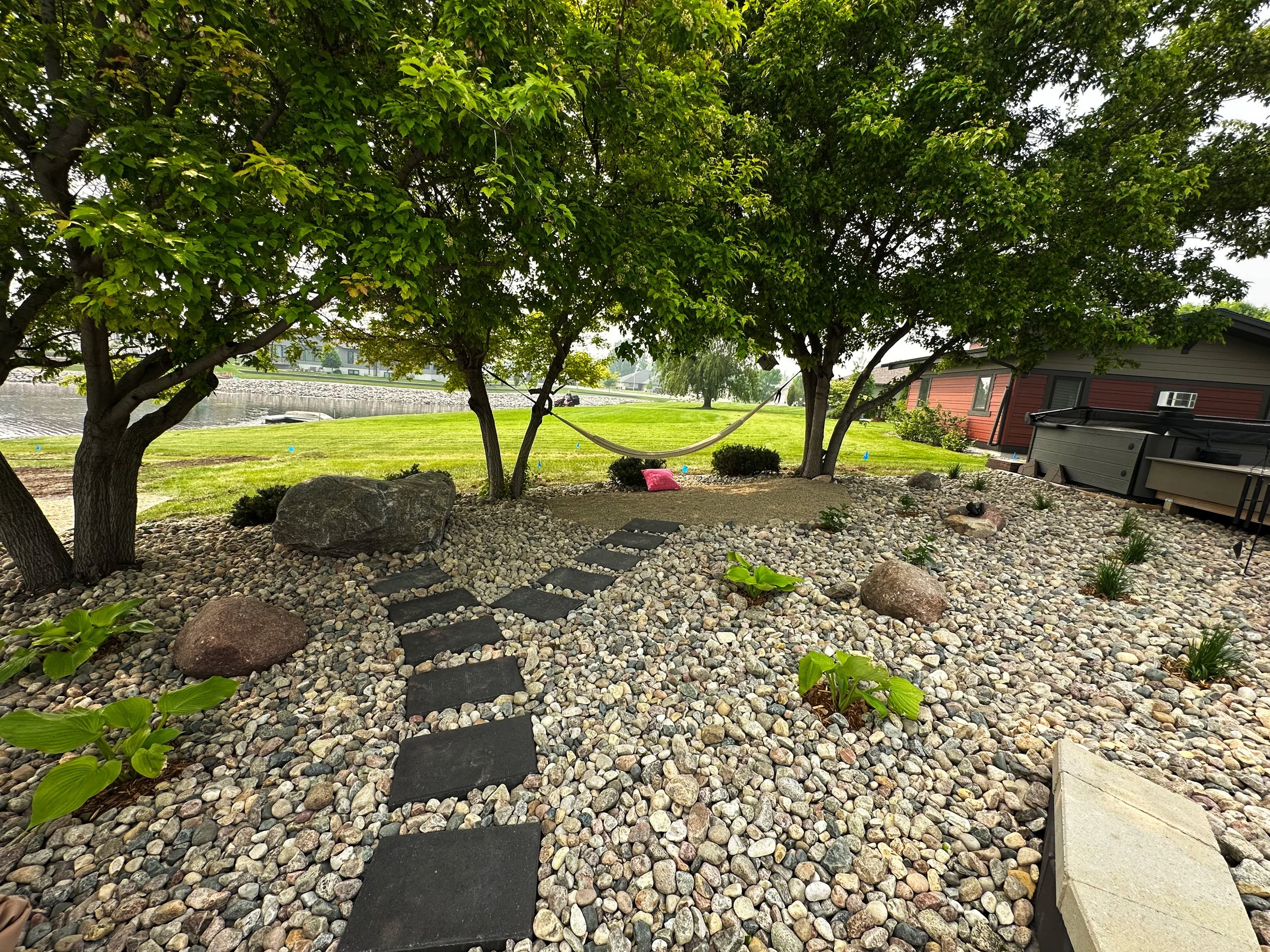 Backyard landscape with three trees, a hammock between two trees, a stone pathway, rocks, small plants, a grassy area, a house, and a body of water in the background.